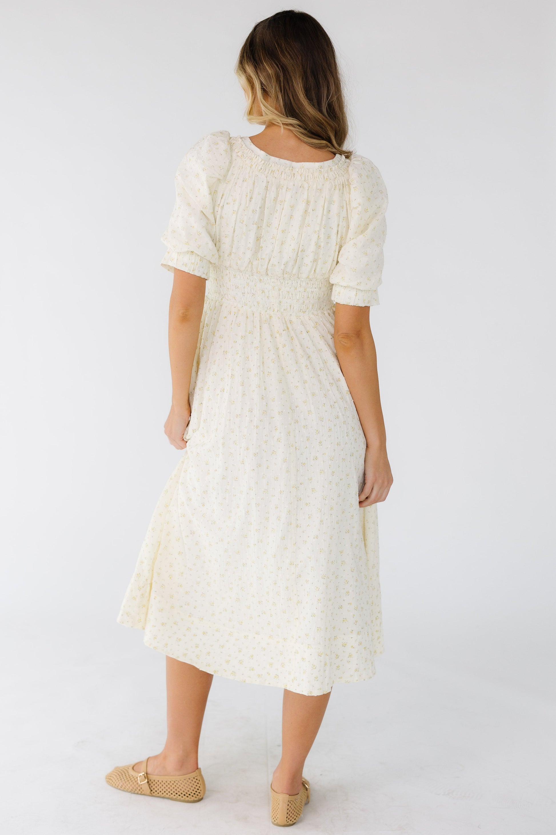 Back view of a woman wearing a cream floral midi dress with s wide waistband and balloon sleeves.