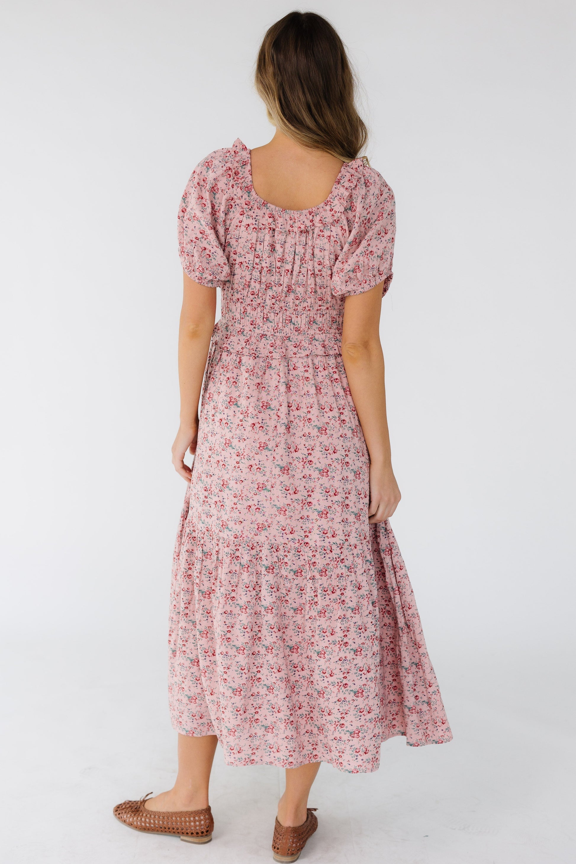 Back view of a woman wearing a pink floral dress with a ruffled scoop neckline, short puff sleeves and a wide waistband and a full ruffled skirt.