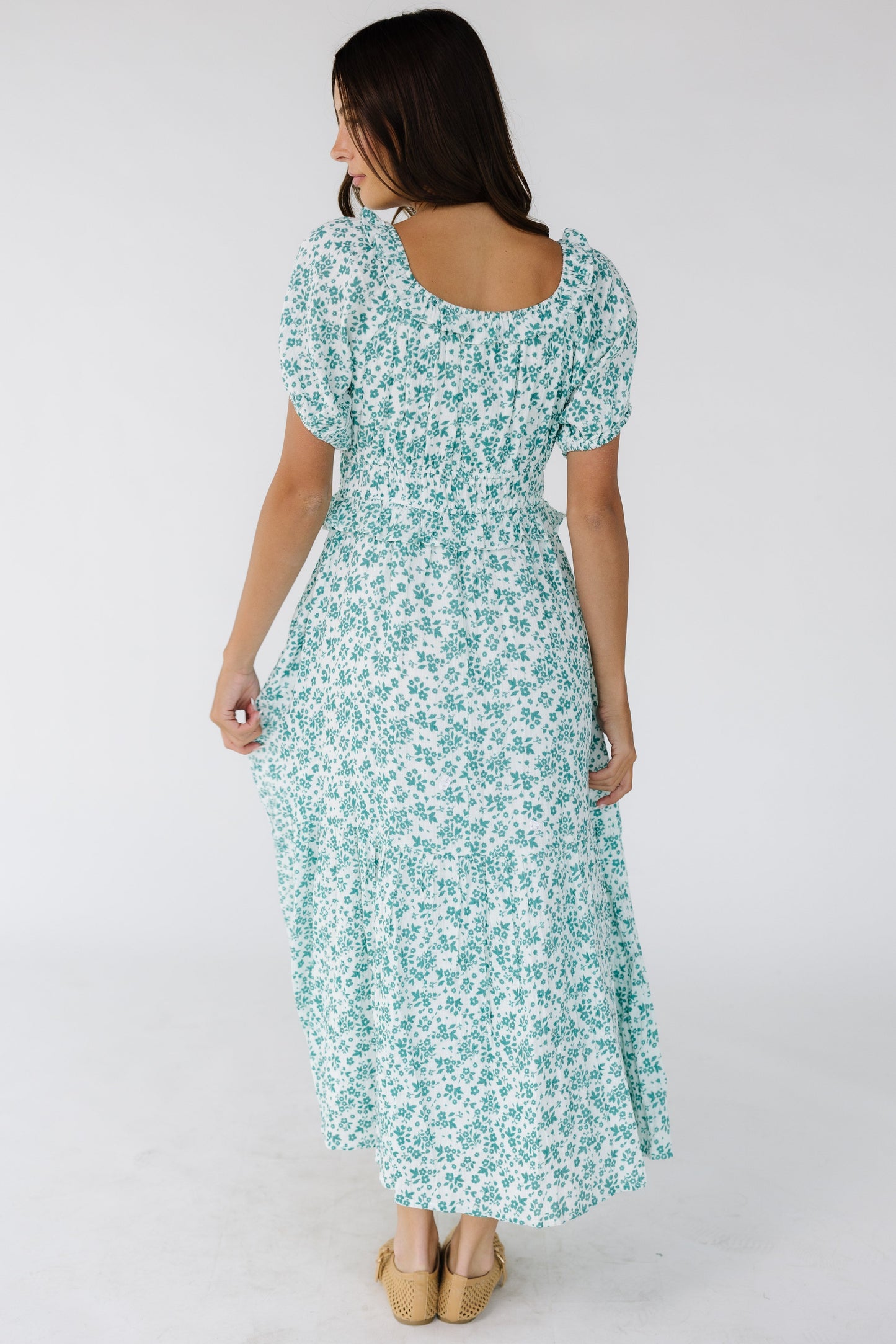 Back view of a woman wearing a white & teal floral print midi dress with short puff sleeves and a wide waistband and a wide bottom ruffle.