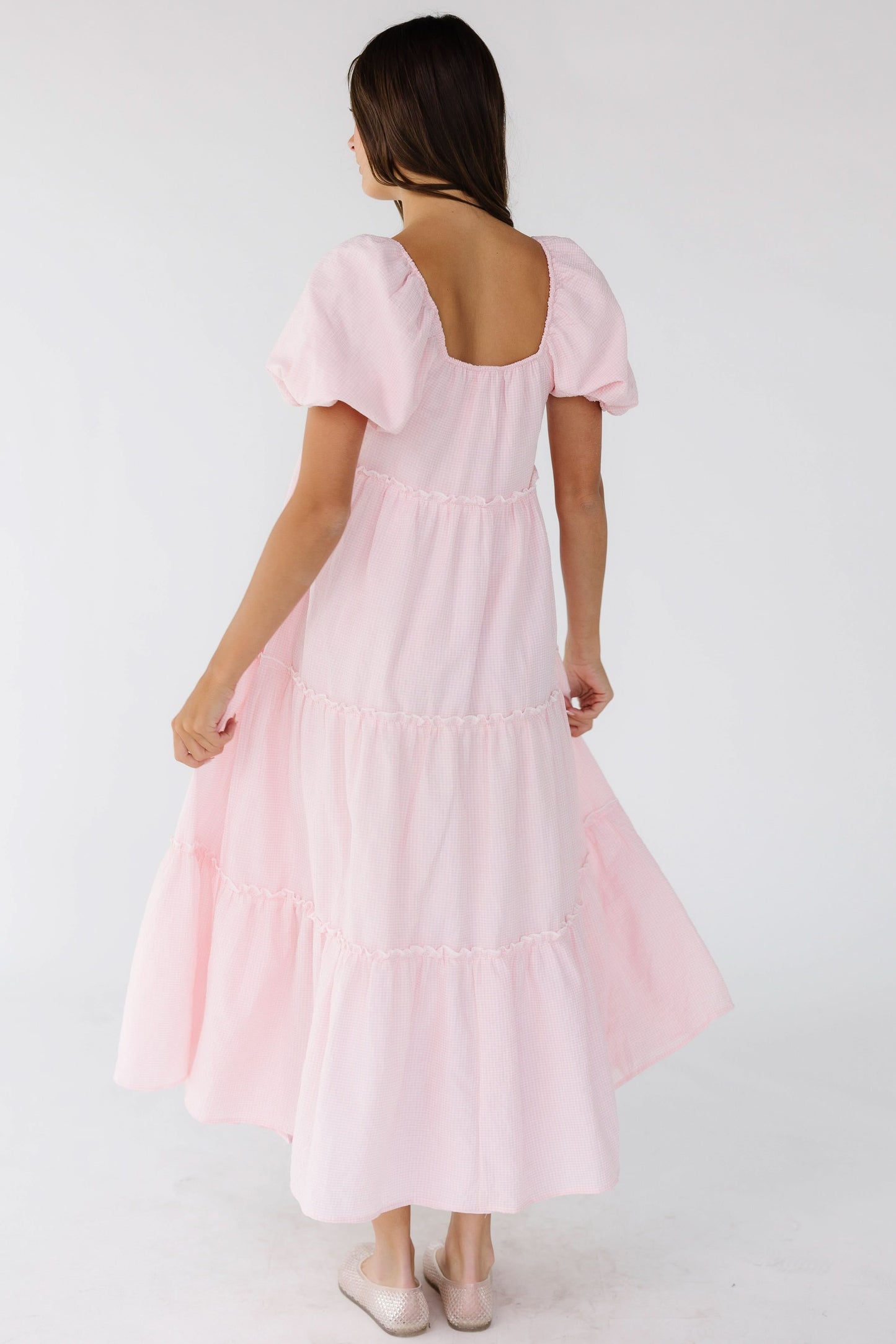 Back view of a woman wearing a pink gingham tiered midi dress with a square back neckline and balloon sleeves.