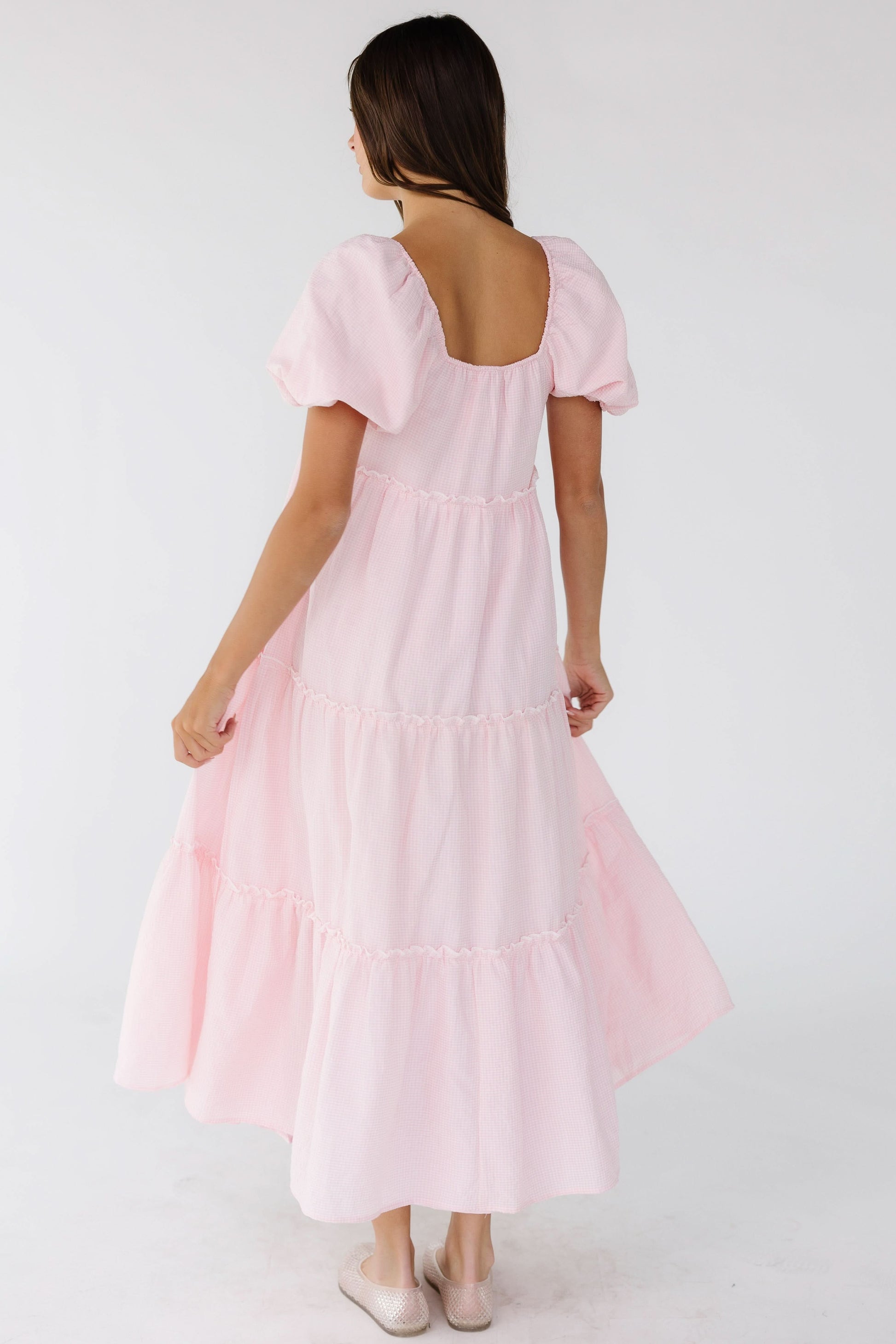 Back view of a woman wearing a pink gingham tiered midi dress with a square back neckline and balloon sleeves.