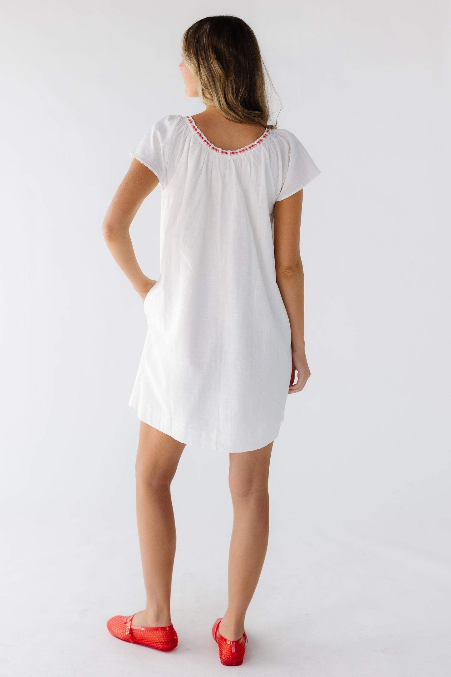 Back view of a woman wearing a short white dress with short sleeves and a red & white gingham trimmed neckline.