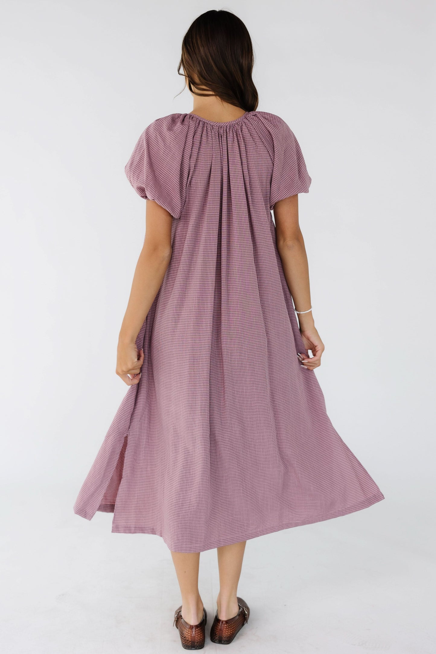 Back view of a woman wearing a loose fitting burgundy & pink gingham midi dress with side slits, and and large puff sleeves.