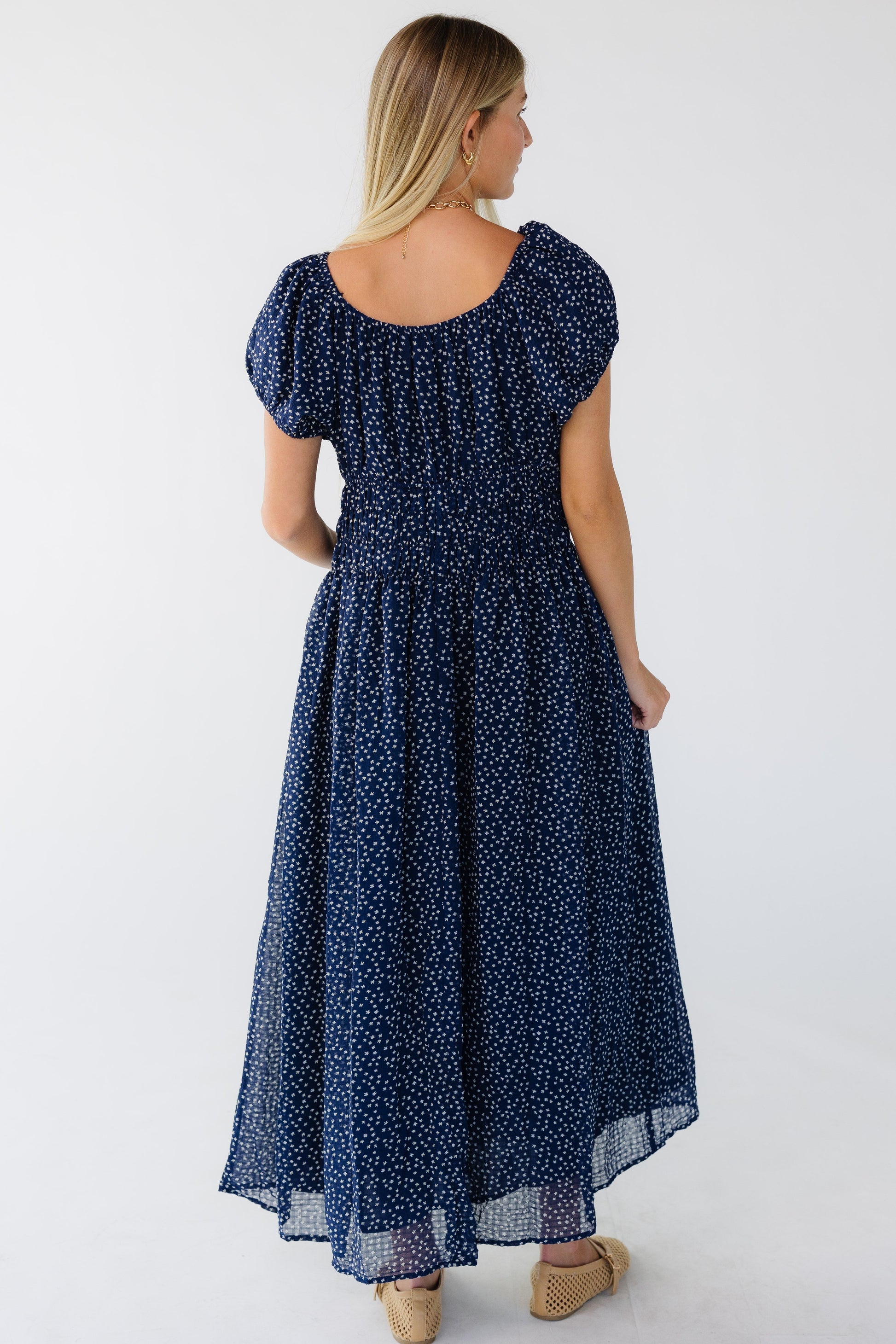 Back view of a woman wearing a navy print midi-maxi dress with a scoop back neckline and short puff sleeves.