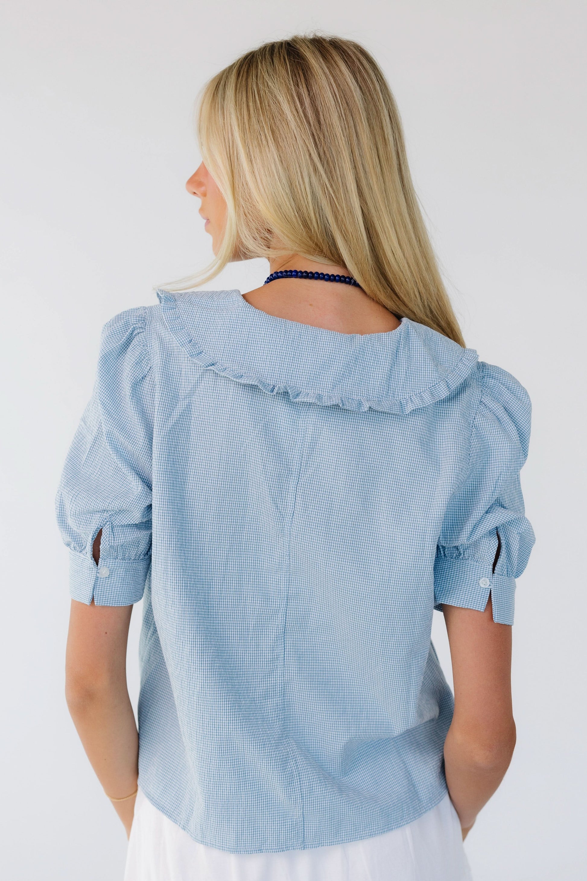 Back view of a woman wearing a light blue gingham, loose fitting blouse with puff sleeves and a ruffle edge collar.