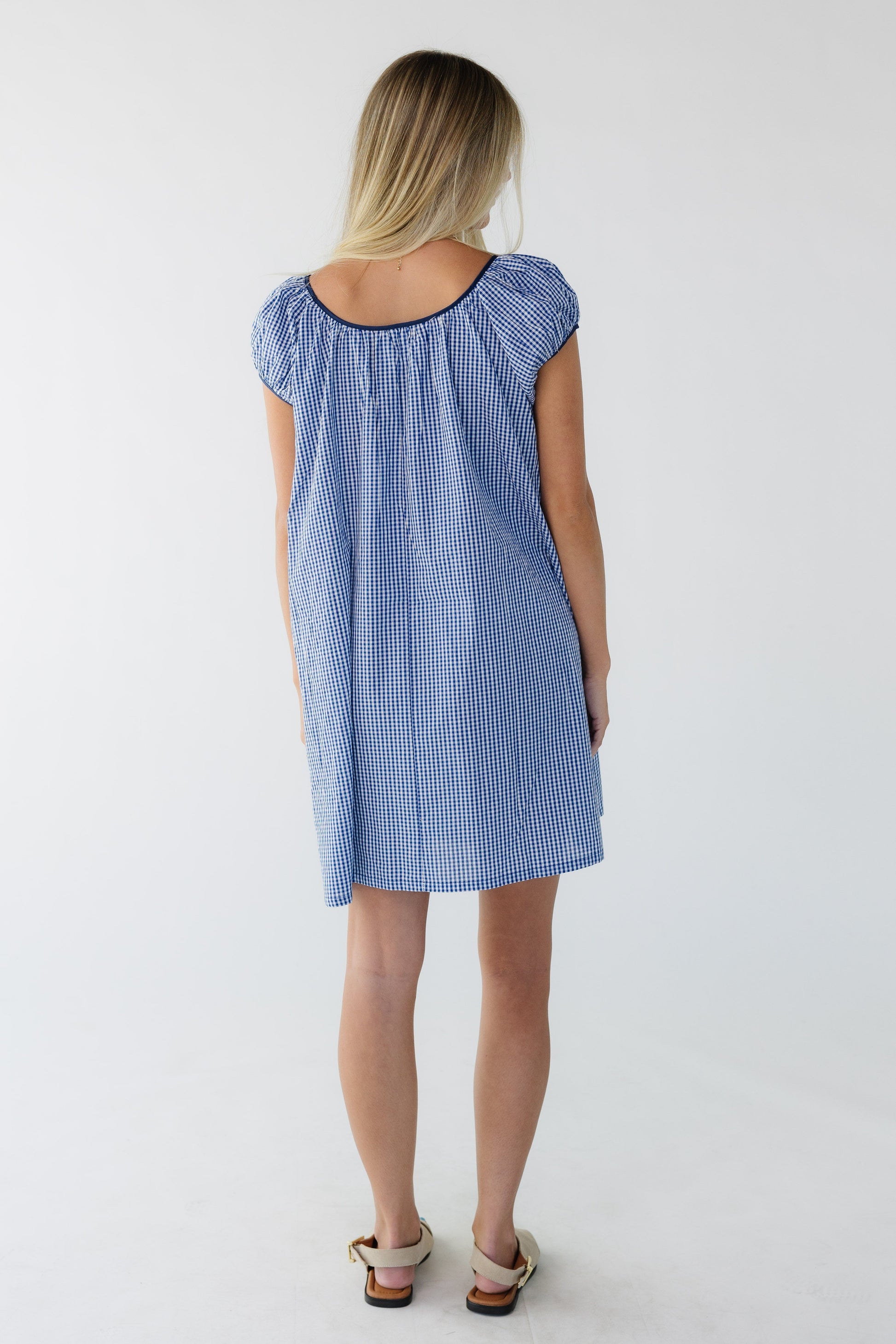 Back view of a woman wearing a navy gingham short dress with short sleeves and a wide scoop neckline.