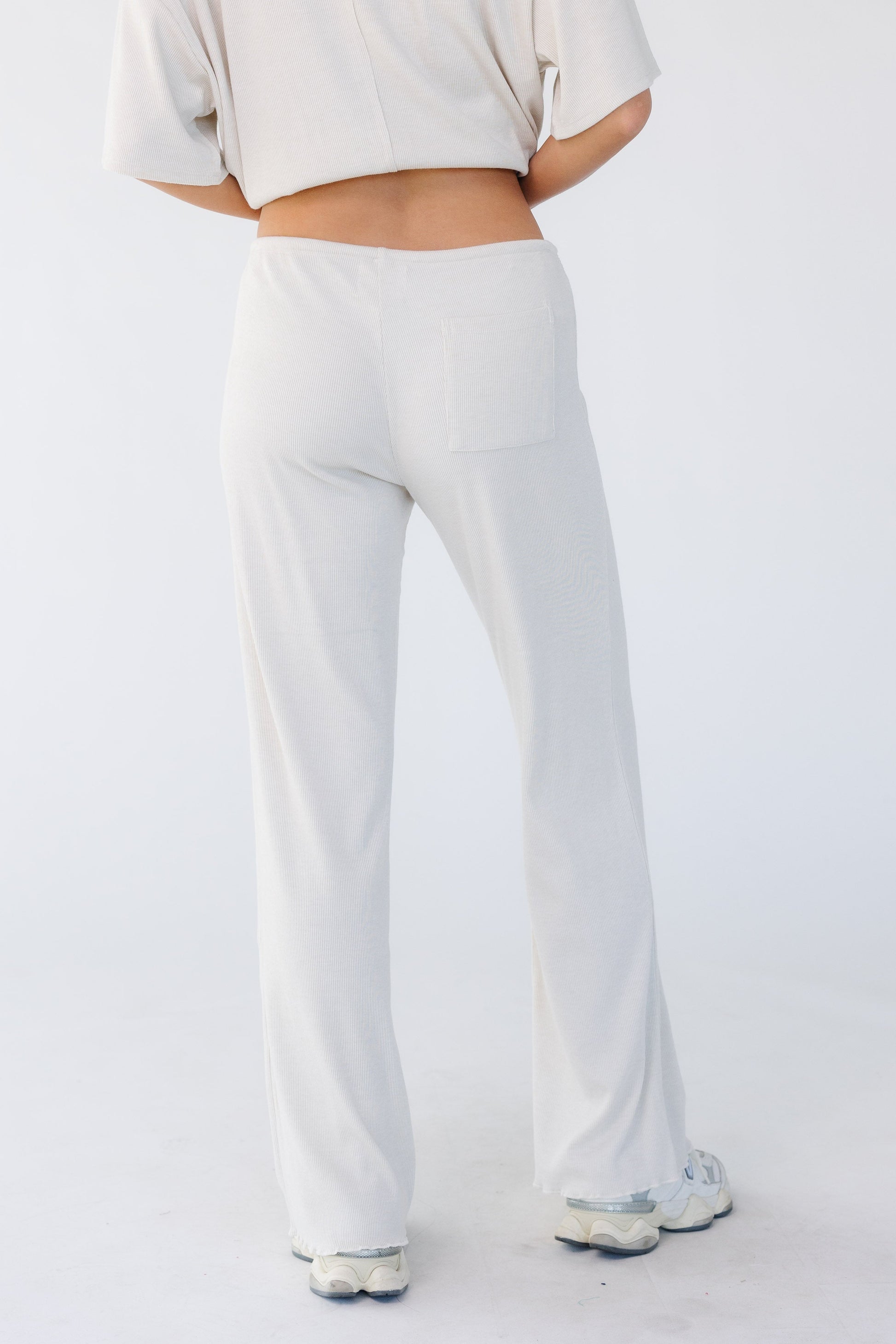 Back view of a young woman wearing a pair of lounge pants that are close fitting through the waist & hip with a wide cut leg in light beige.