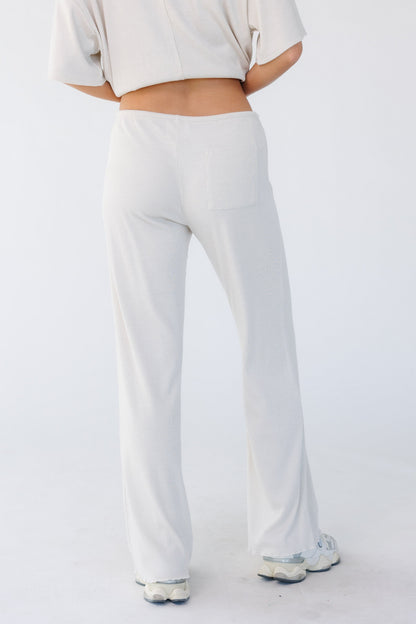 Back view of a young woman wearing a pair of lounge pants that are close fitting through the waist & hip with a wide cut leg in light beige.
