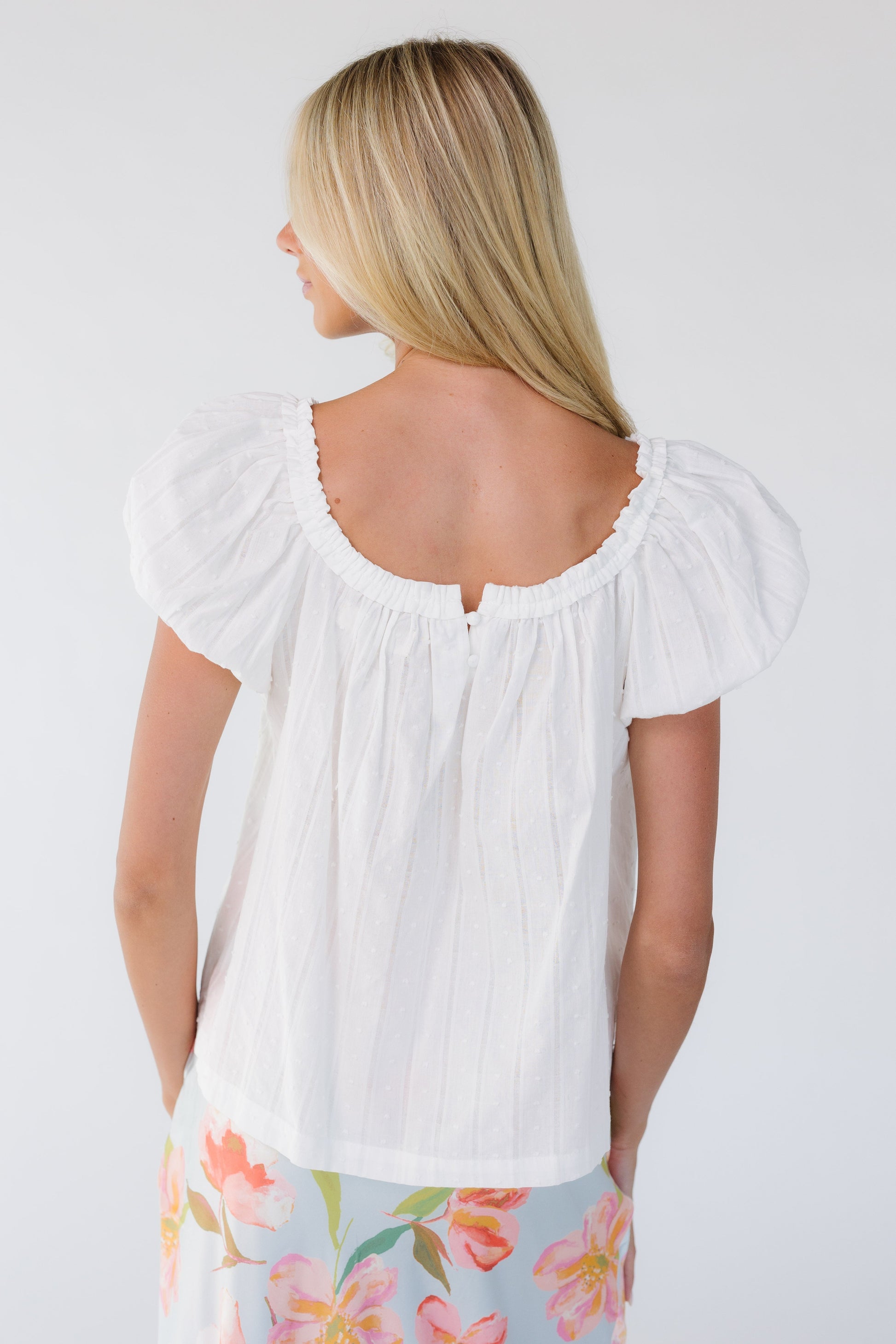 Back view of a young woman wearing a white Swiss dot top with a square neckline and short puff sleeves.
