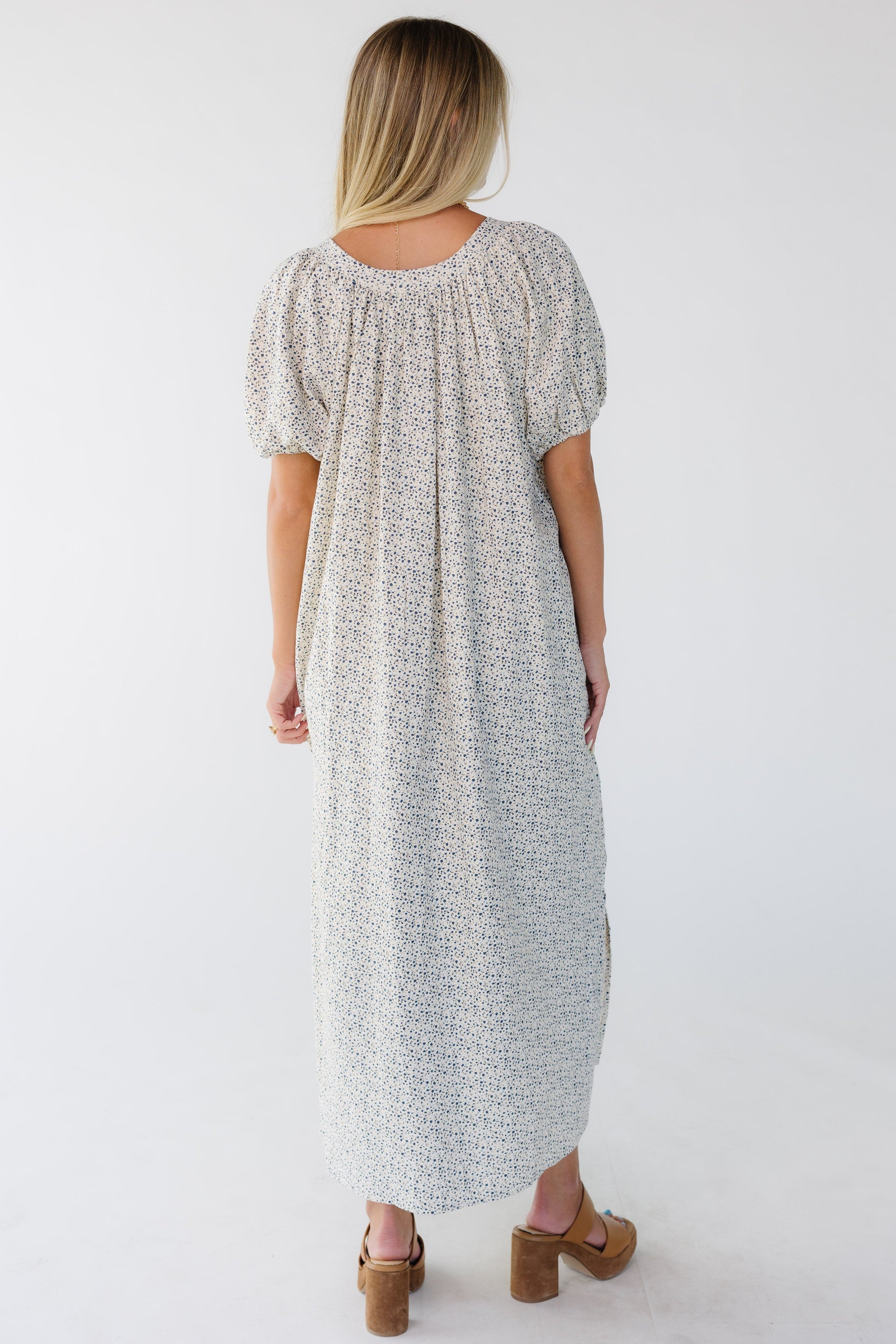 Back view of a woman wearing a cream & blue floral print midi-maxi dress with a back scoop neckline and balloon sleeves.