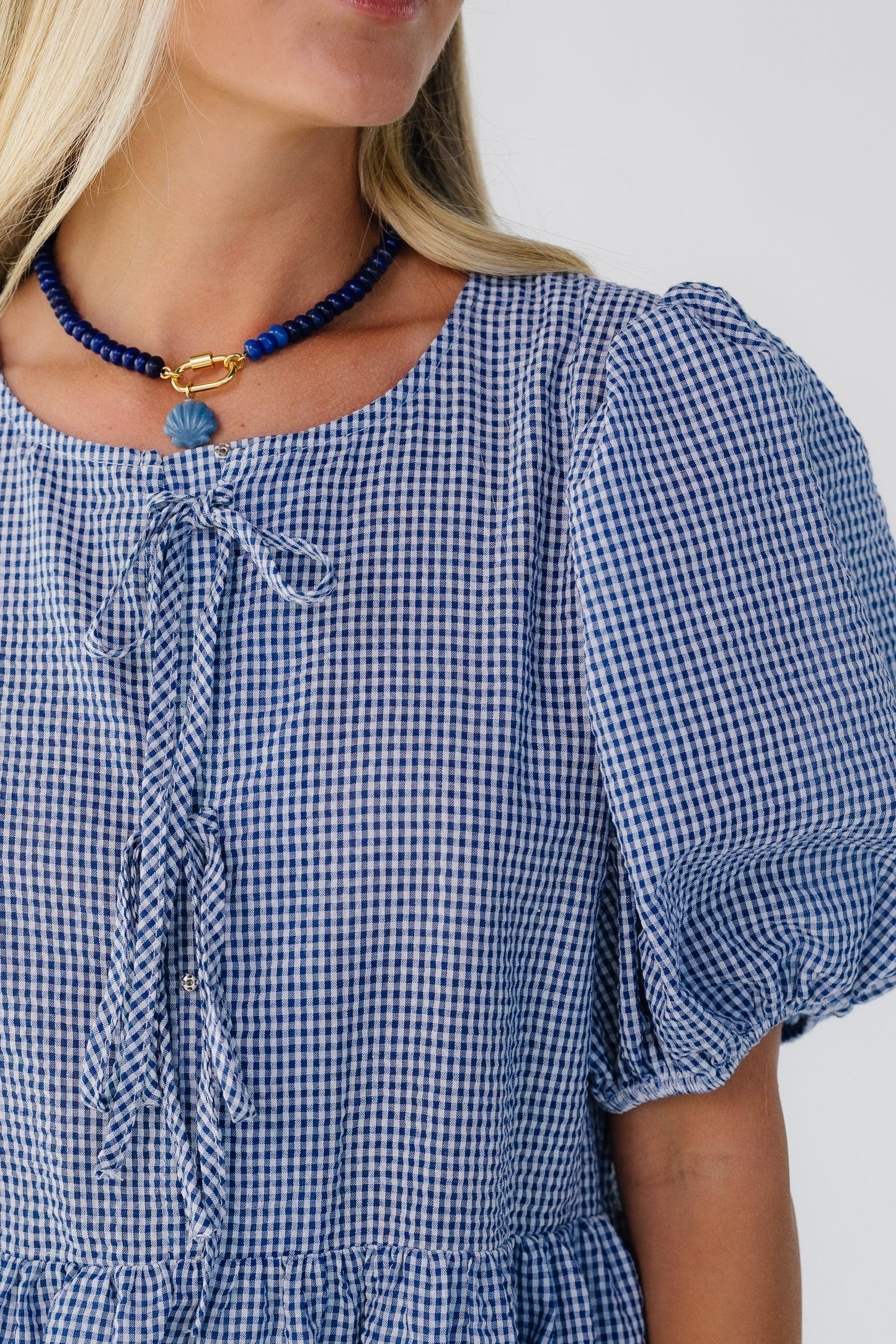 Close view of a woman wearing a blue & white gingham dress with a round neckline, large balloon sleeves and 3 bow closure.