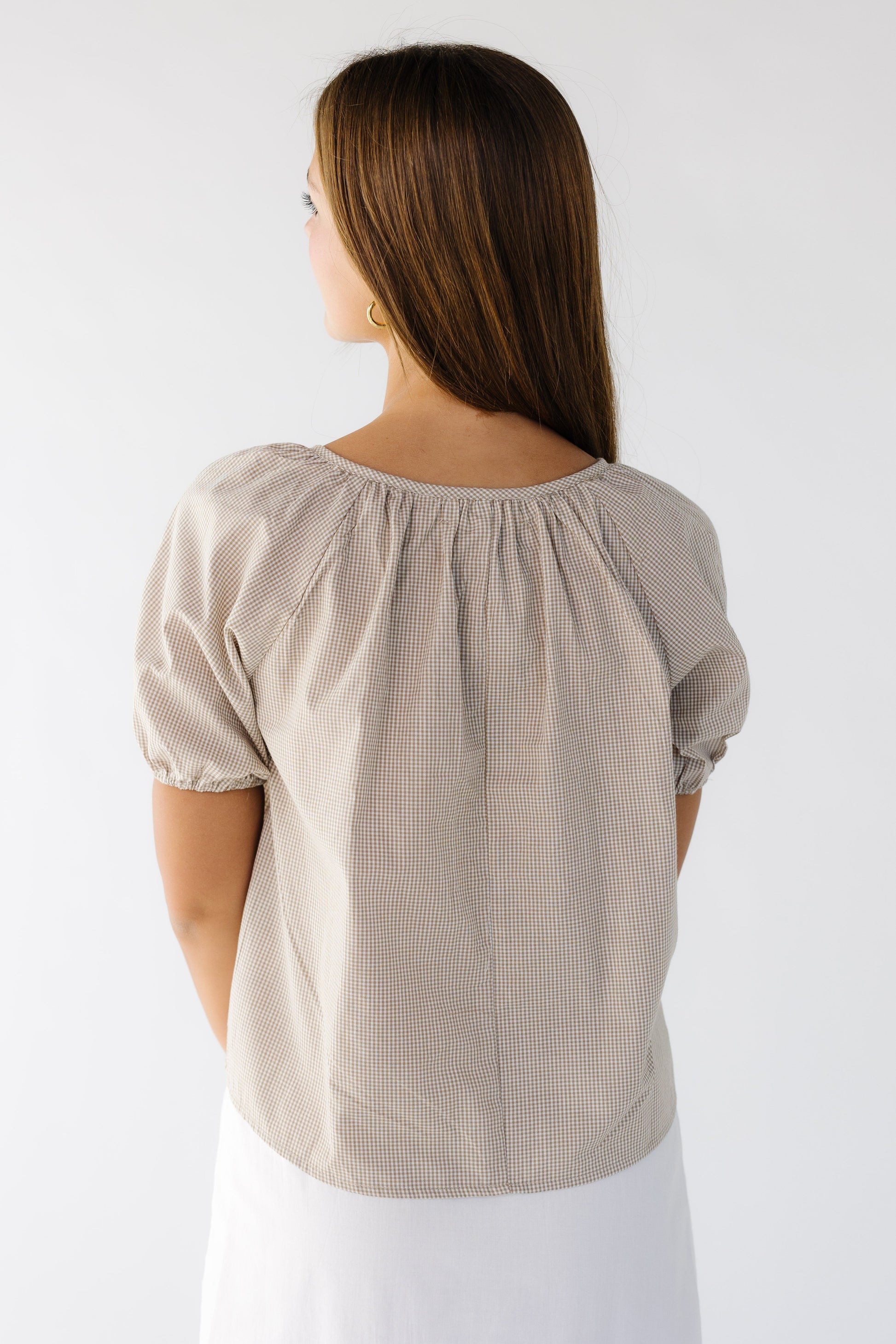 Back view of a woman wearing a loose fitting brown gingham blouse with short puff sleeves.