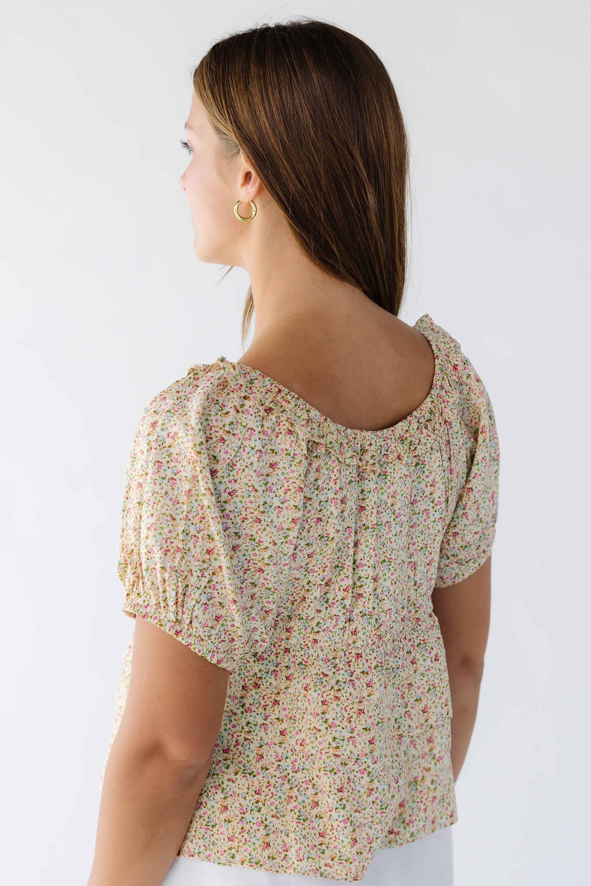 Back view of a woman wearing a yellow floral top with short puff sleeves and a round ruffled neckline.