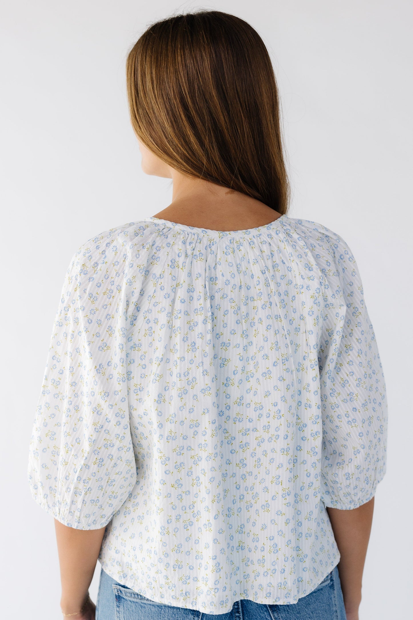 Back view of a young woman wearing a white with blue floral print swing top with elbow length sleeves and a round neckline.
