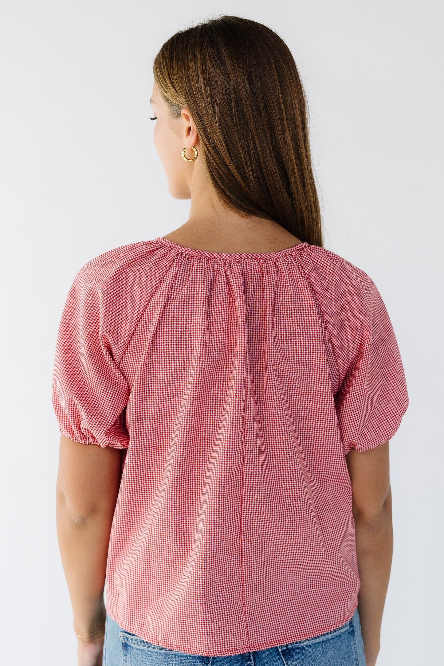 Back view of a woman wearing a loose fitting red gingham blouse  with short puff sleeves. 