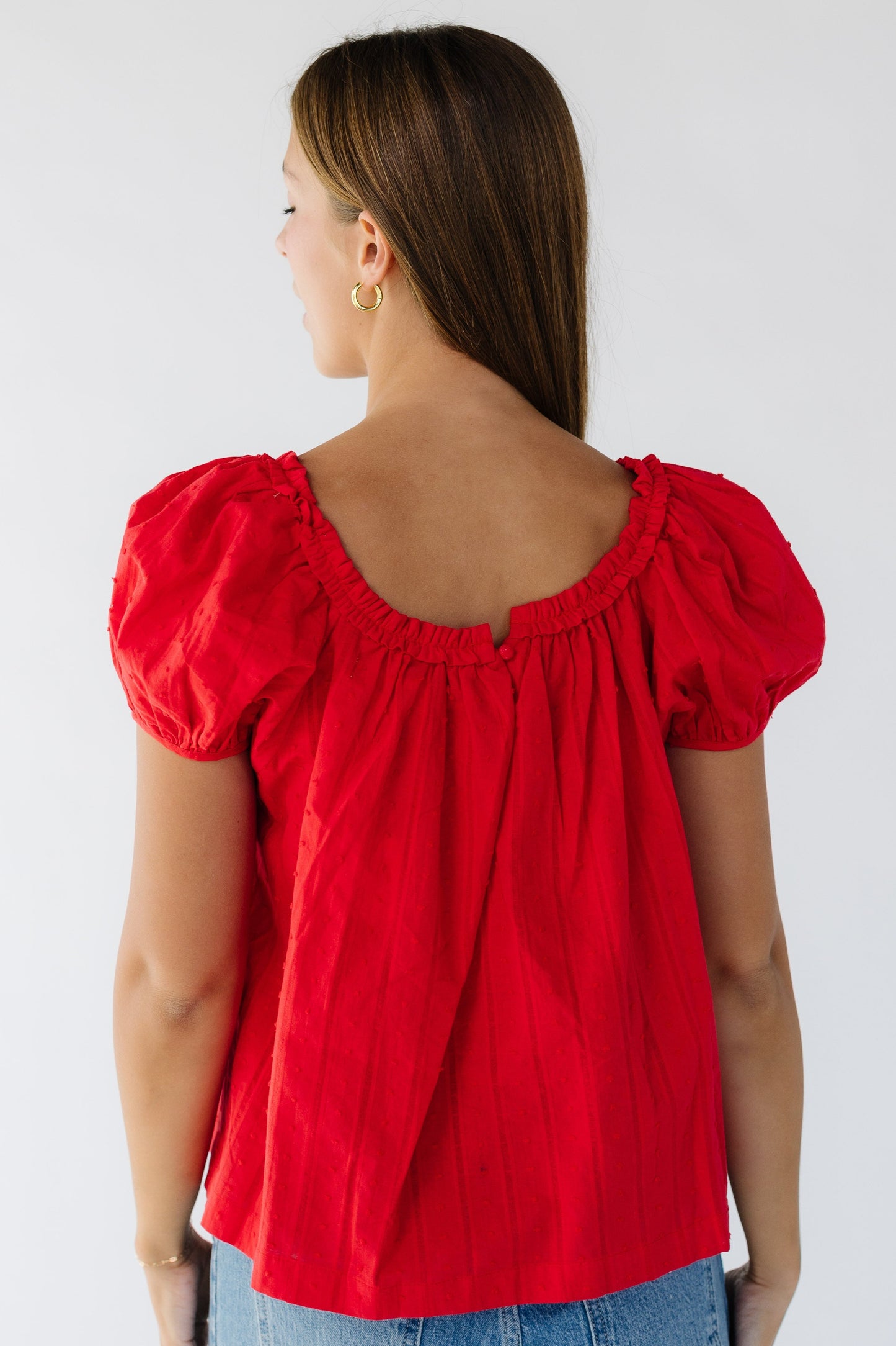 Back view of a young woman wearing a loose fitting red blouse with a ruffled square neckline and short puff sleeves.