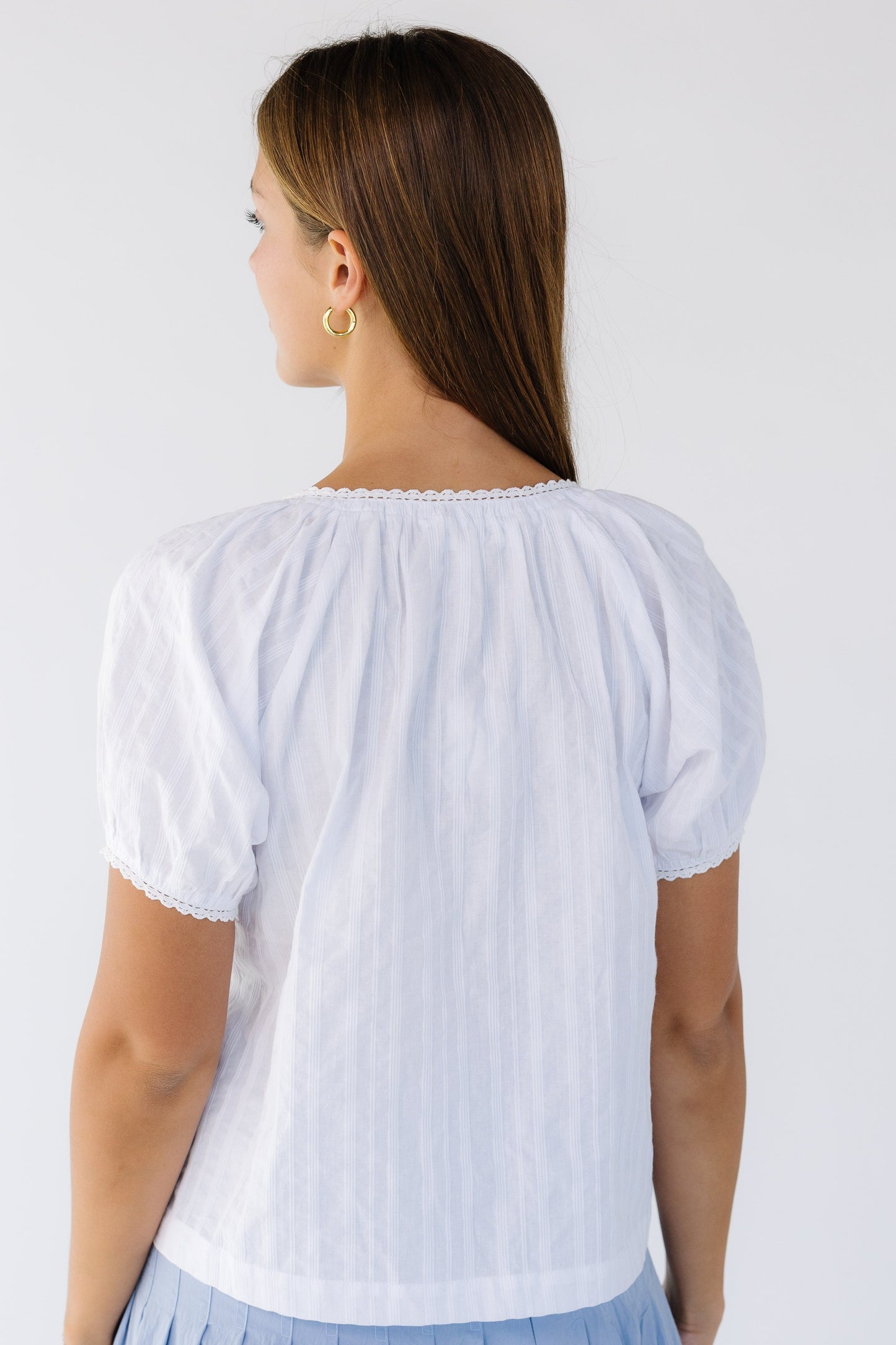 Back view of a woman wearing a white loose fitting blouse with a lace trimmed neckline and lace trimmed puff sleeves.