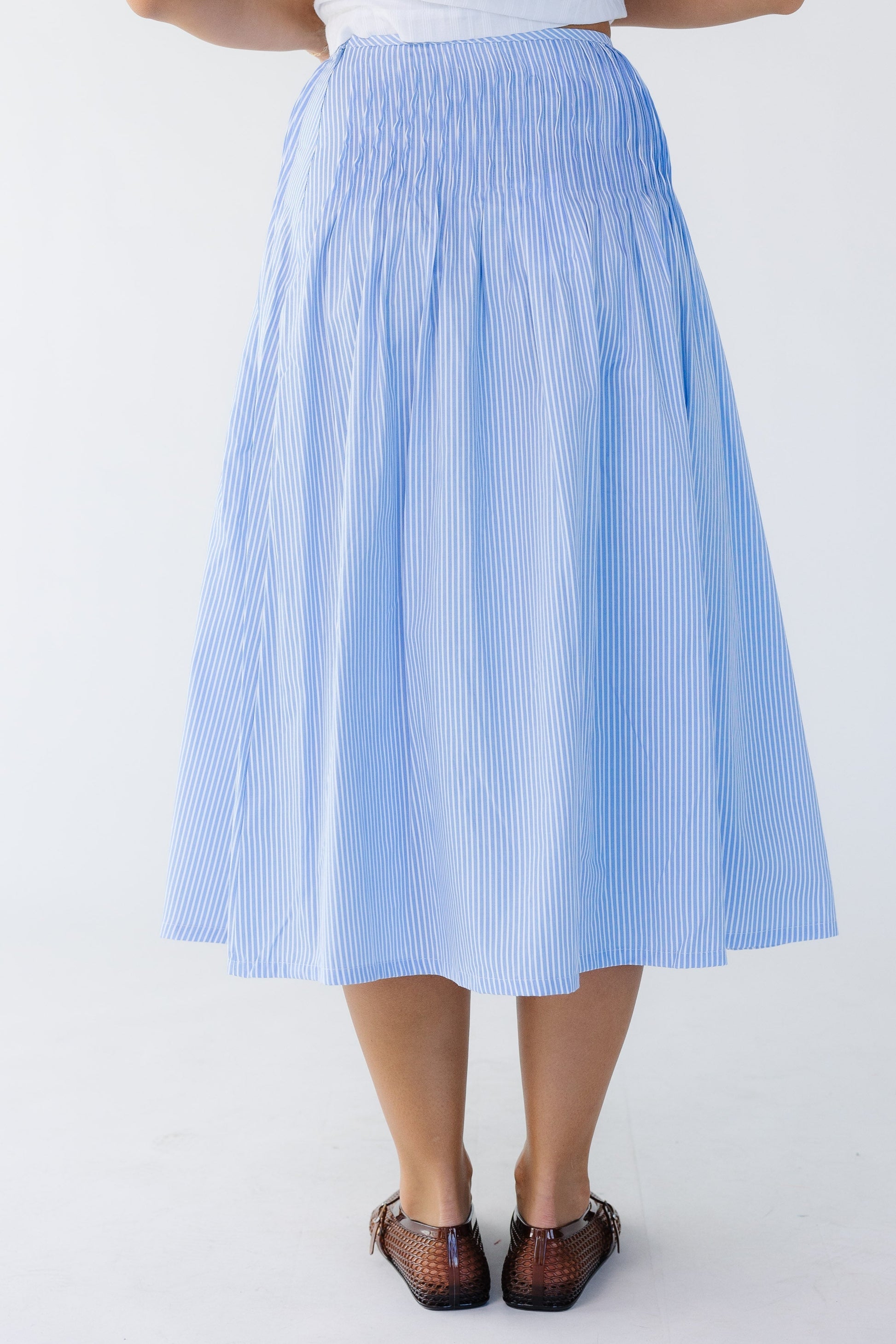 Back view of a woman wearing a blue and white stripe midi skirt with sewn down pleats all around the hips.