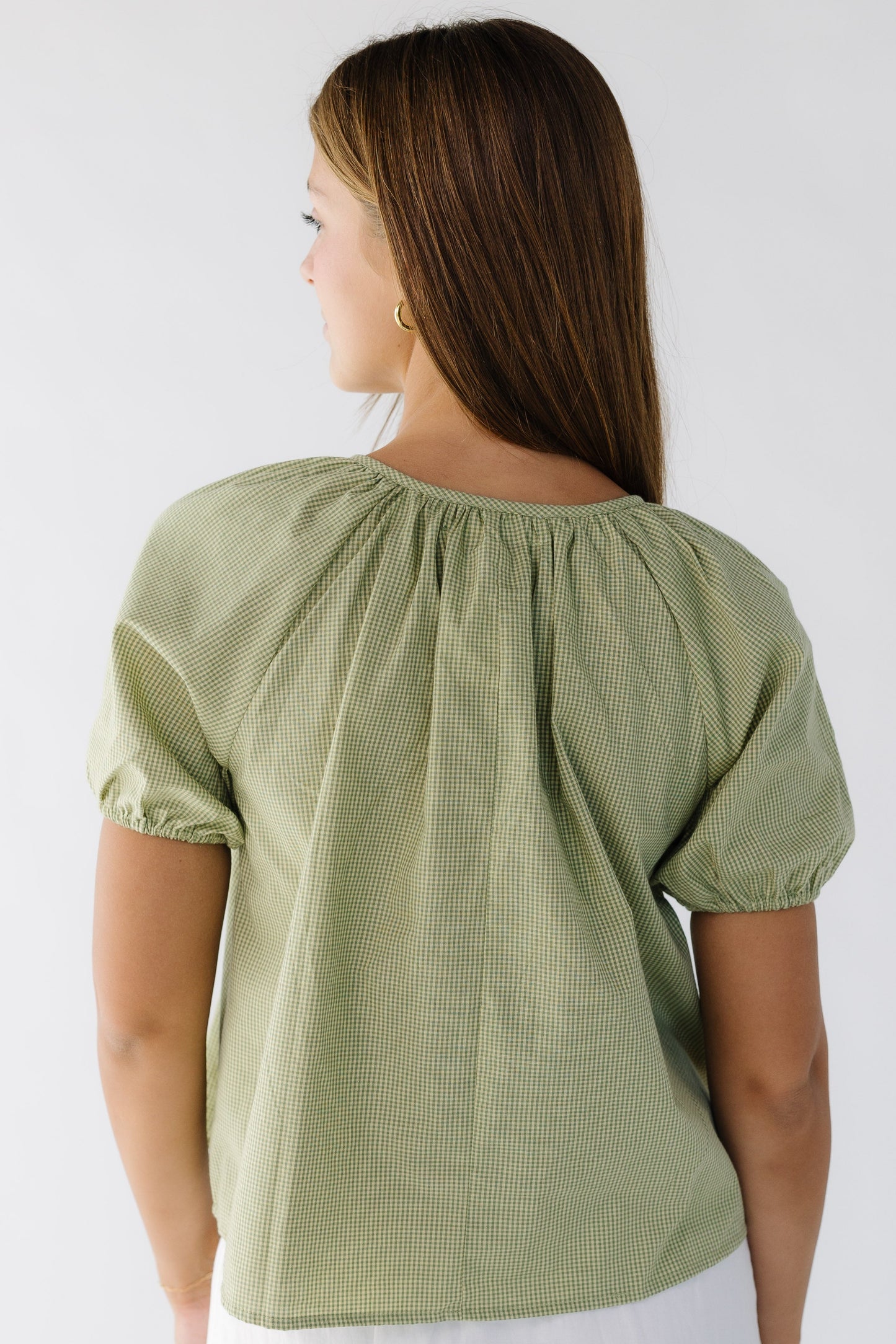 Back view of a woman wearing a green gingham loose fitting blouse with short puff sleeves.
