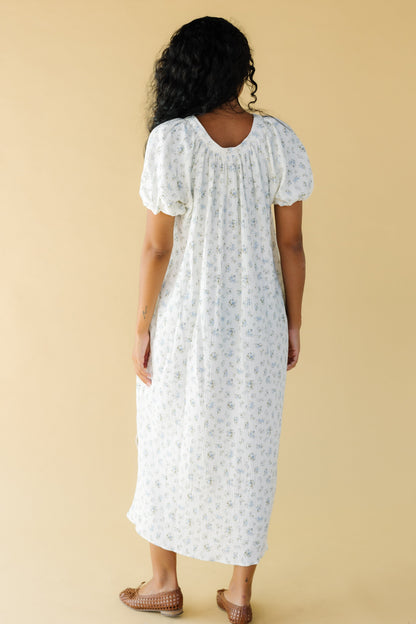 Back view of a woman wearing a white & blue floral midi dress with a scoop neckline and balloon sleeves.