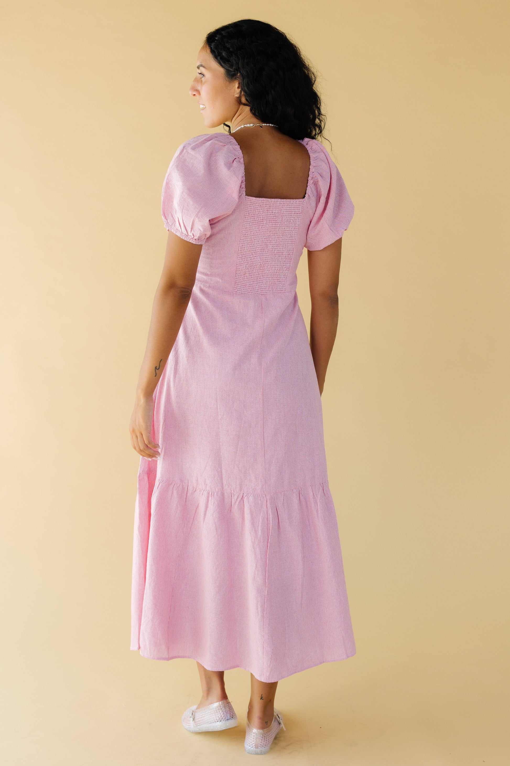 Back view of a woman wearing a red & white stripe midi-maxi dress with an elastic smocked back bodice panel, square back neckline large puff sleeves  and a wide bottom ruffle.