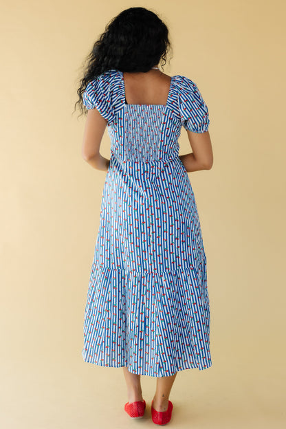 Back view of a young woman wearing a blue & white-cherry print midi-maxi dress with and elastic smocked back bodice panel, large puff sleeves and a full skirt with a wide bottom ruffle.