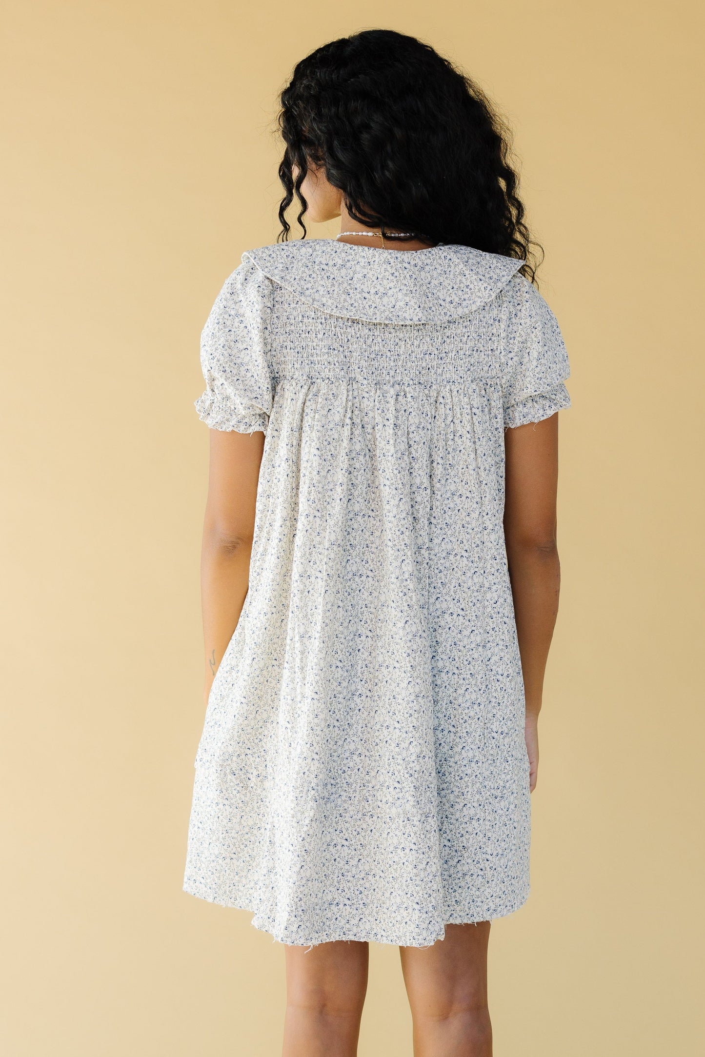 Back view of a young woman wearing a above knee length dress with a wide collar, smocked back yoke and short puff sleeves.