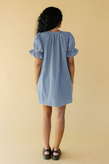 Back view of a young woman wearing a navy & white gingham short dress with ruffle hemmed puff sleeves.