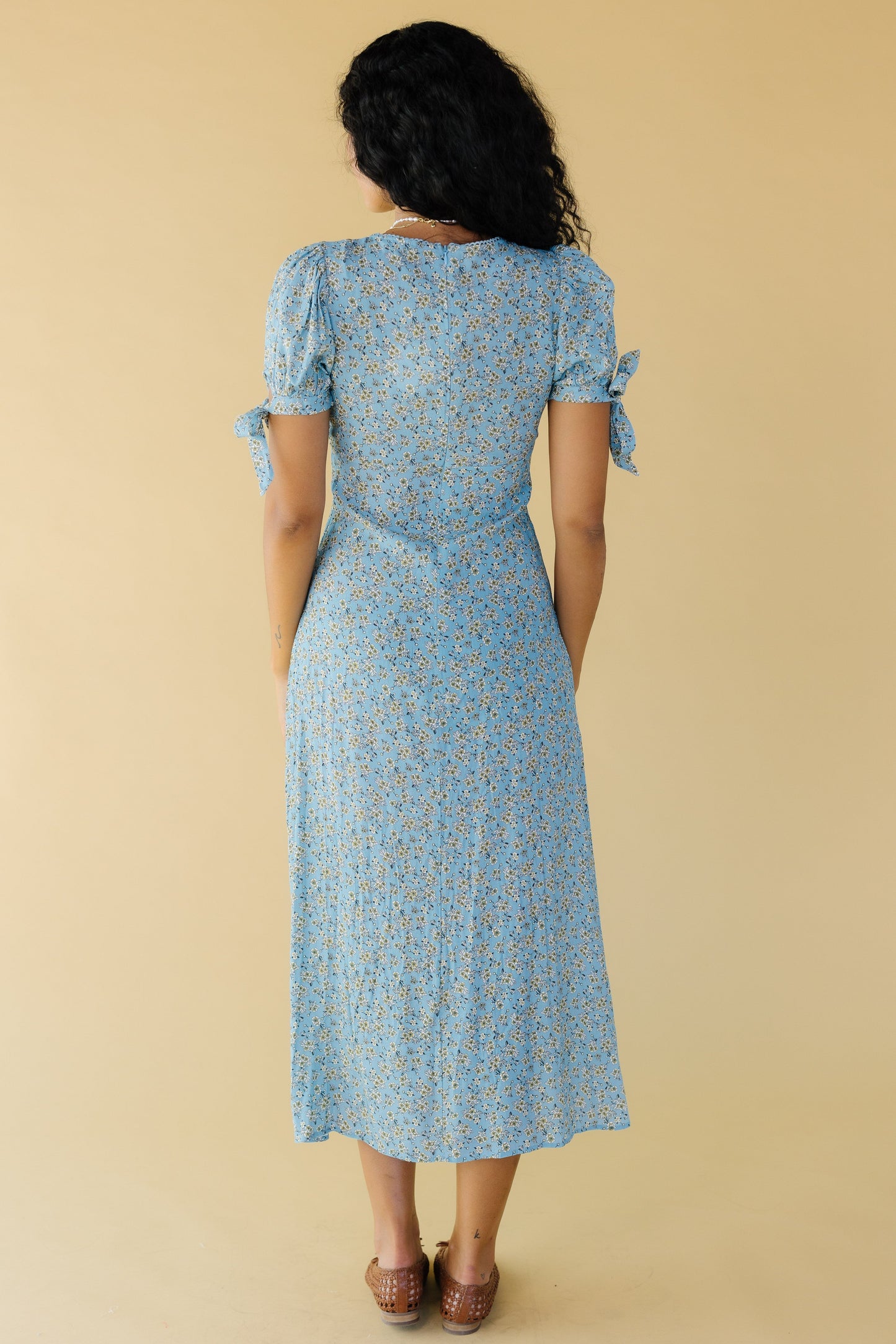 Back view of a woman wearing a light blue floral midi dress with puff sleeves and tied cuffs.