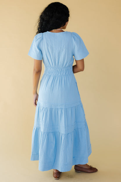 Back view of a woman wearing a light blue midi-maxi dress with short sleeves and a wide 3 band elastic waistband.