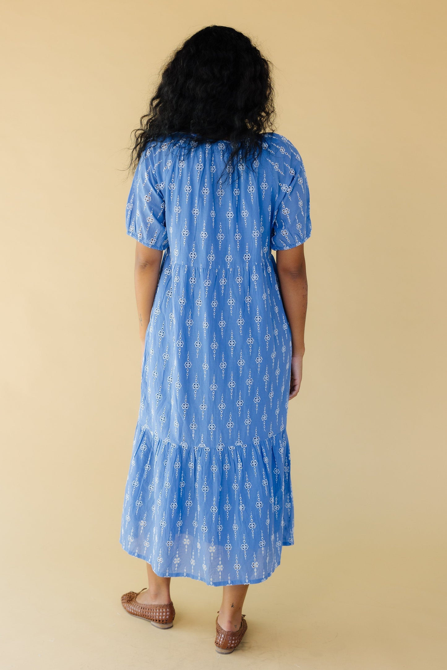 Back view of a woman wearing a blue midi dress with white eyelet embroidery, short puff sleeves and a wide bottom ruffle.