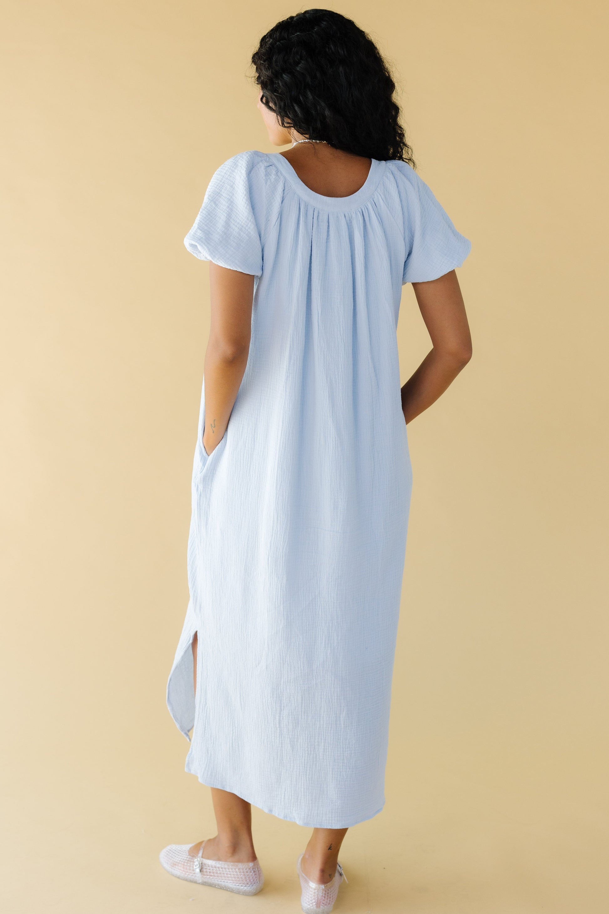 Back view of a woman wearing a light blue midi dress with side slits, pockets and large puffy sleeves.