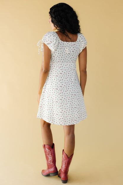 Back view of a young woman wearing a cream & red floral short dress with a square back neckline and short puff sleeves.