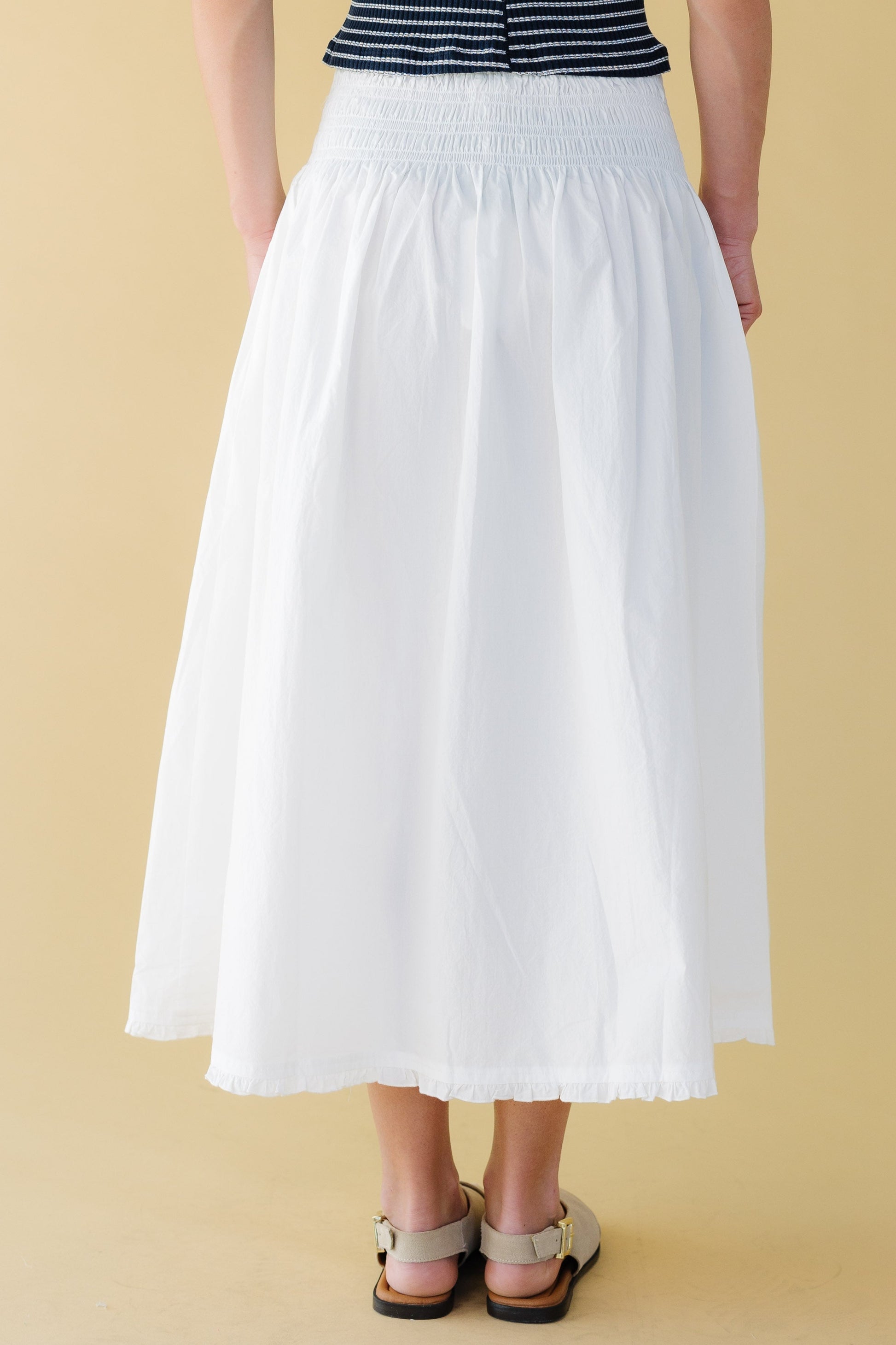 Back view of a woman wearing a white gathered skirt with a wide elastic smocked waist and a narrow ruffle hem.