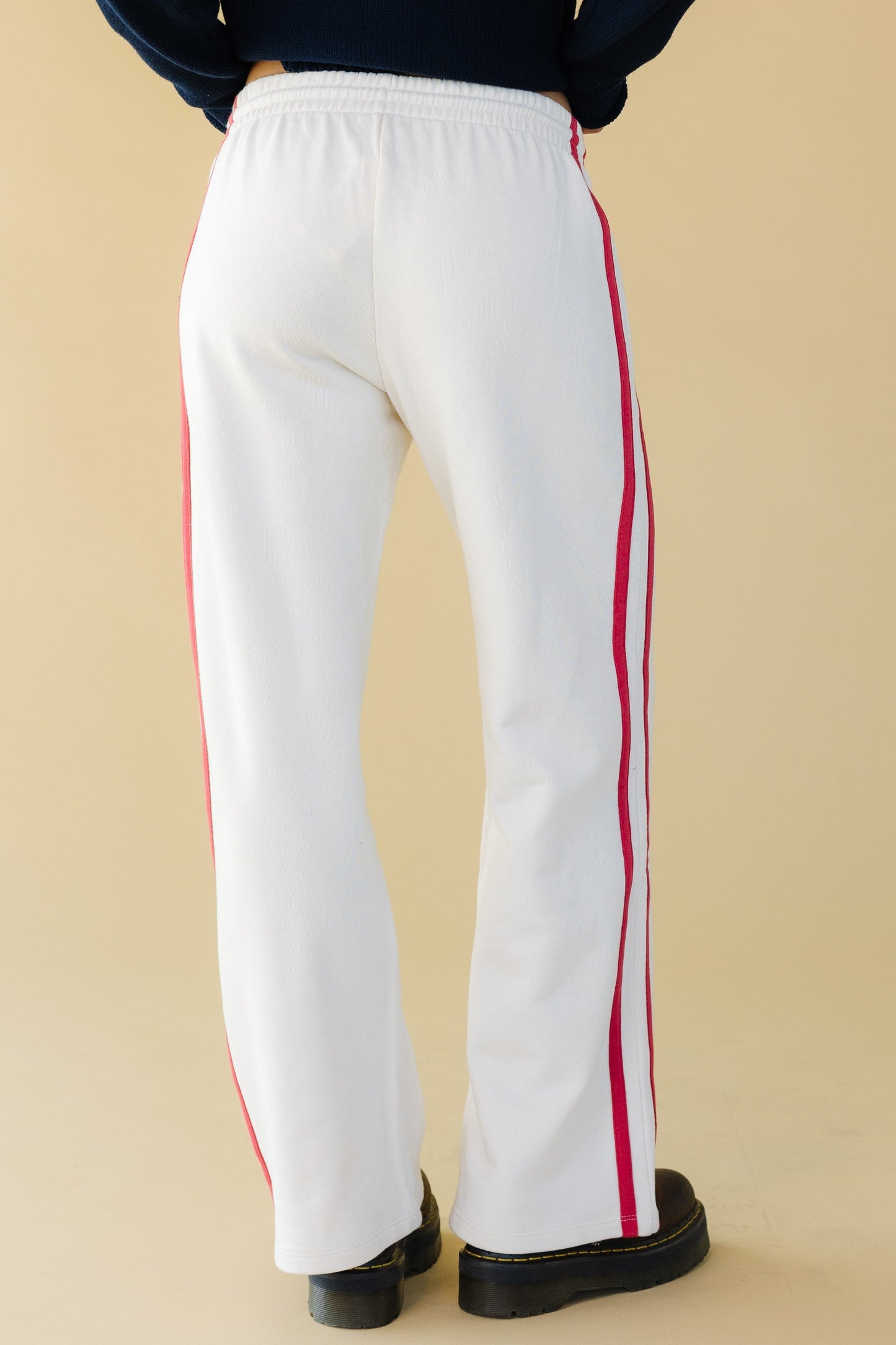 Back view of a young woman wearing white lounge pants with an elastic waist and red stripes down the outside leg.