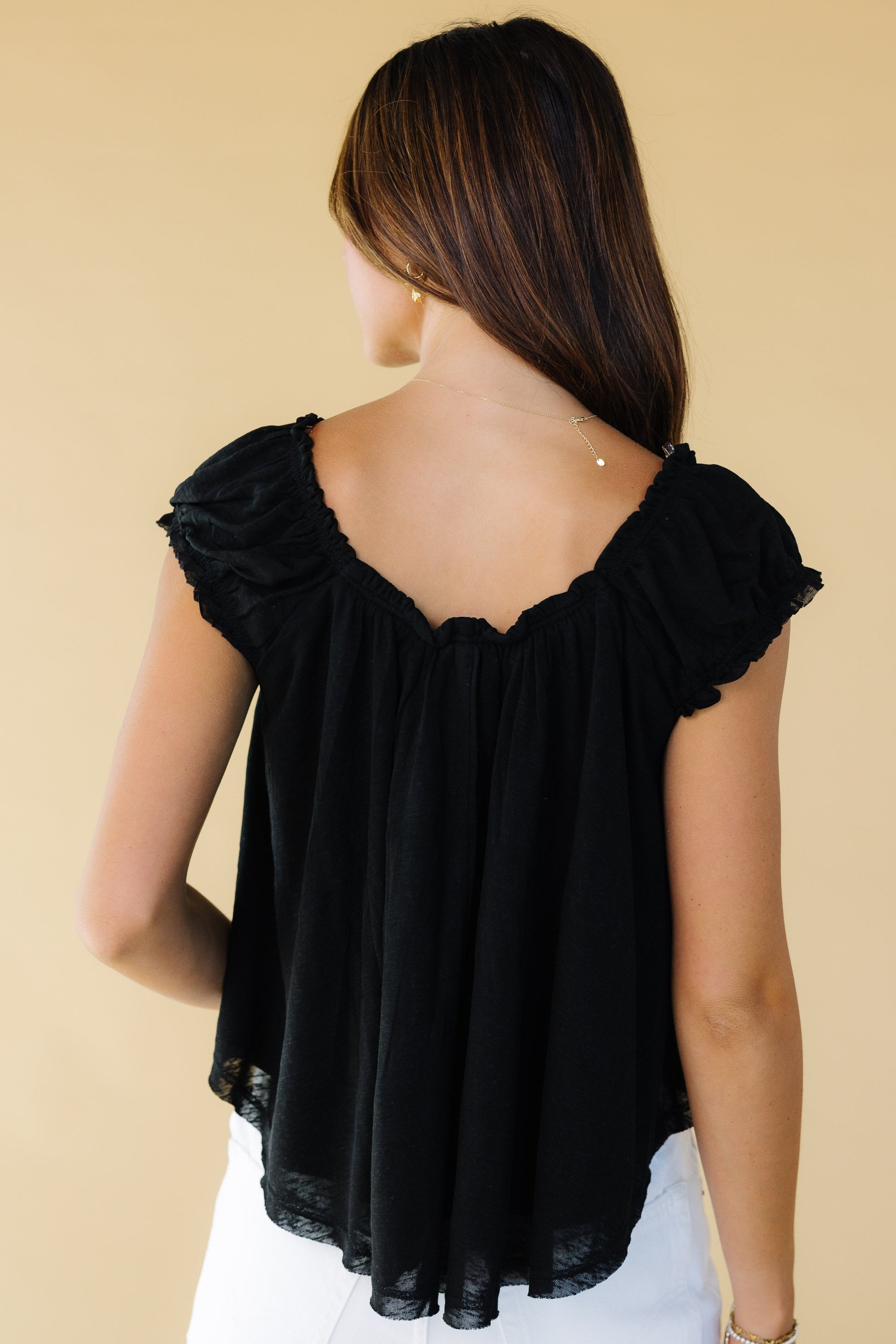 Back view of a woman wearing a loose fitting black blouse with a scoop back neckline and gathered cap sleeves.
