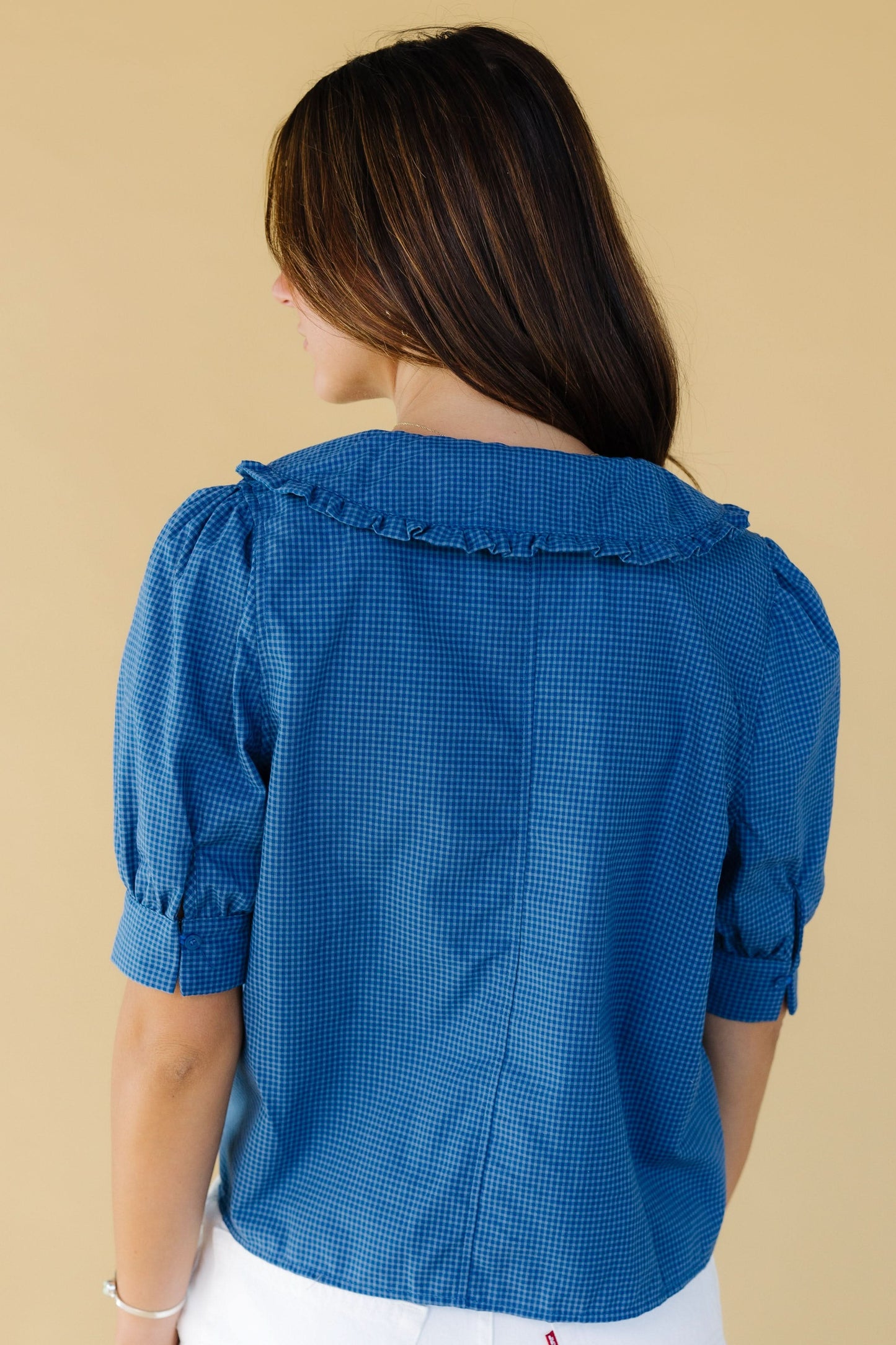 Back view of a young woman wearing a loose fitting blue gingham blouse with cuffed puff sleeves and a wide ruffle edge collar.