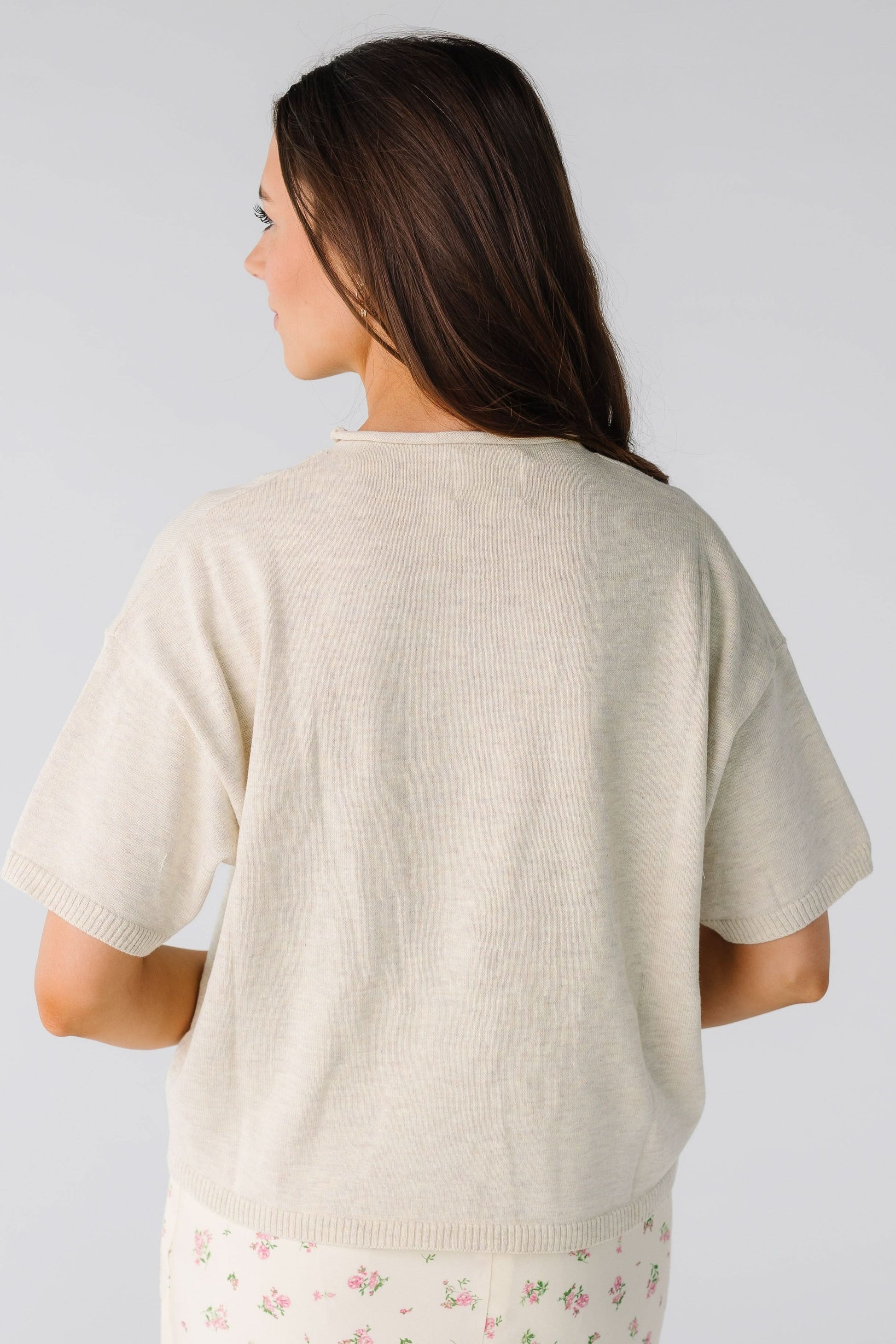 Back view of a young woman wearing a loose fitting sweater ith short sleeves in oatmeal-beige.