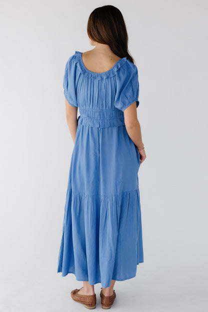 Back view of a young woman wearing a blue polkadot dress with a side bottom ruffle, closer fitting waist and a ruffled scoop back neckline.