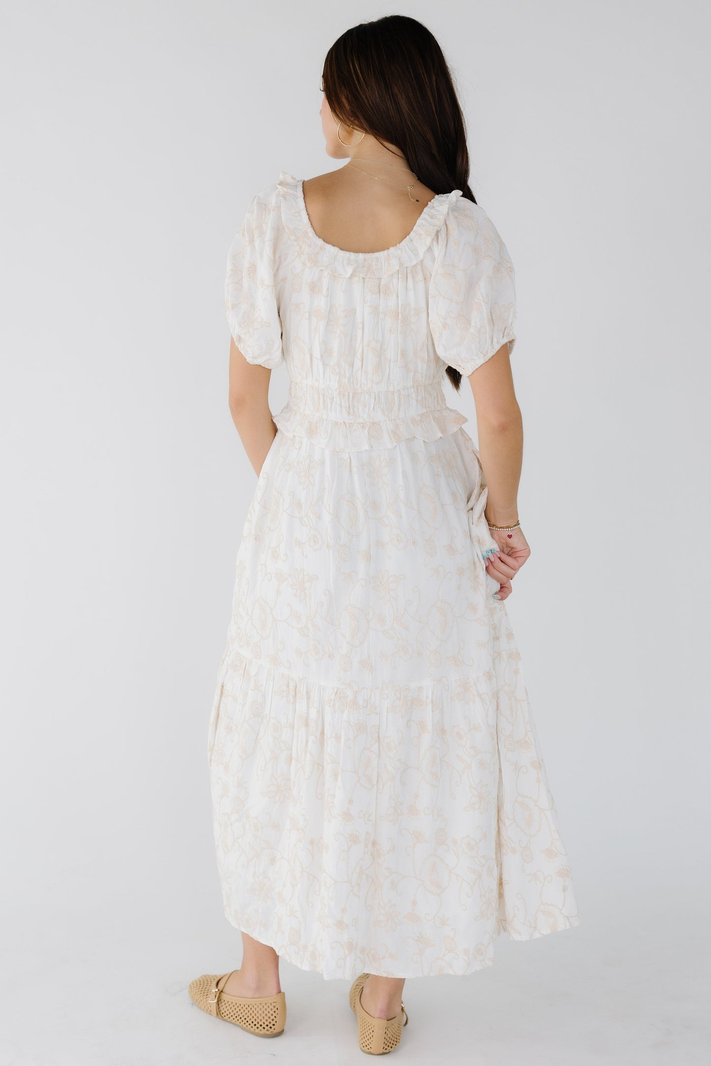 Back view of a young woman wearing a white embroidered midi-maxi dress with a wide waist and short puff sleeves.