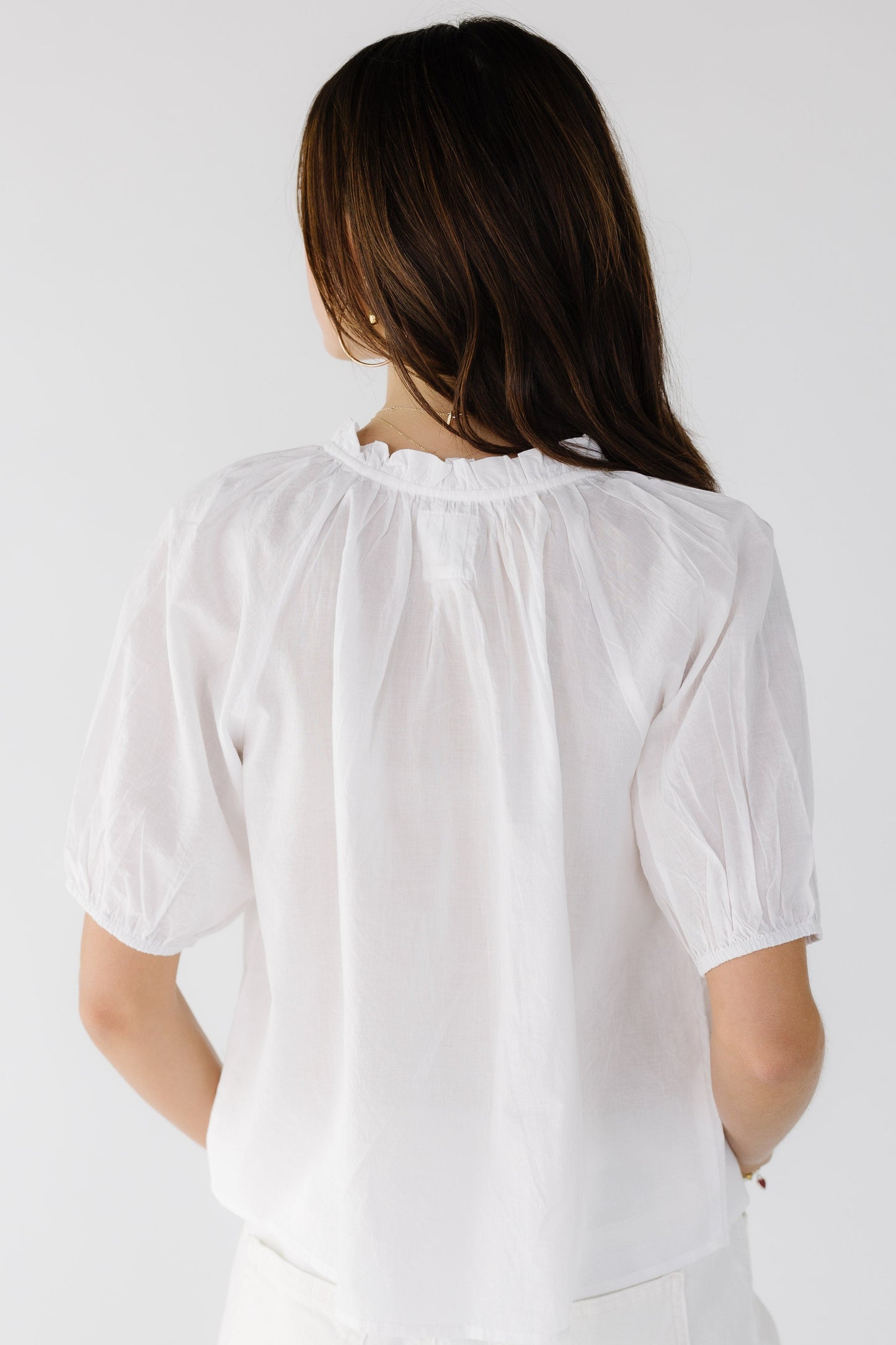 Back view of a young woman wearing a white loose fitting blouse with short gathered sleeves and a ruffled collar.