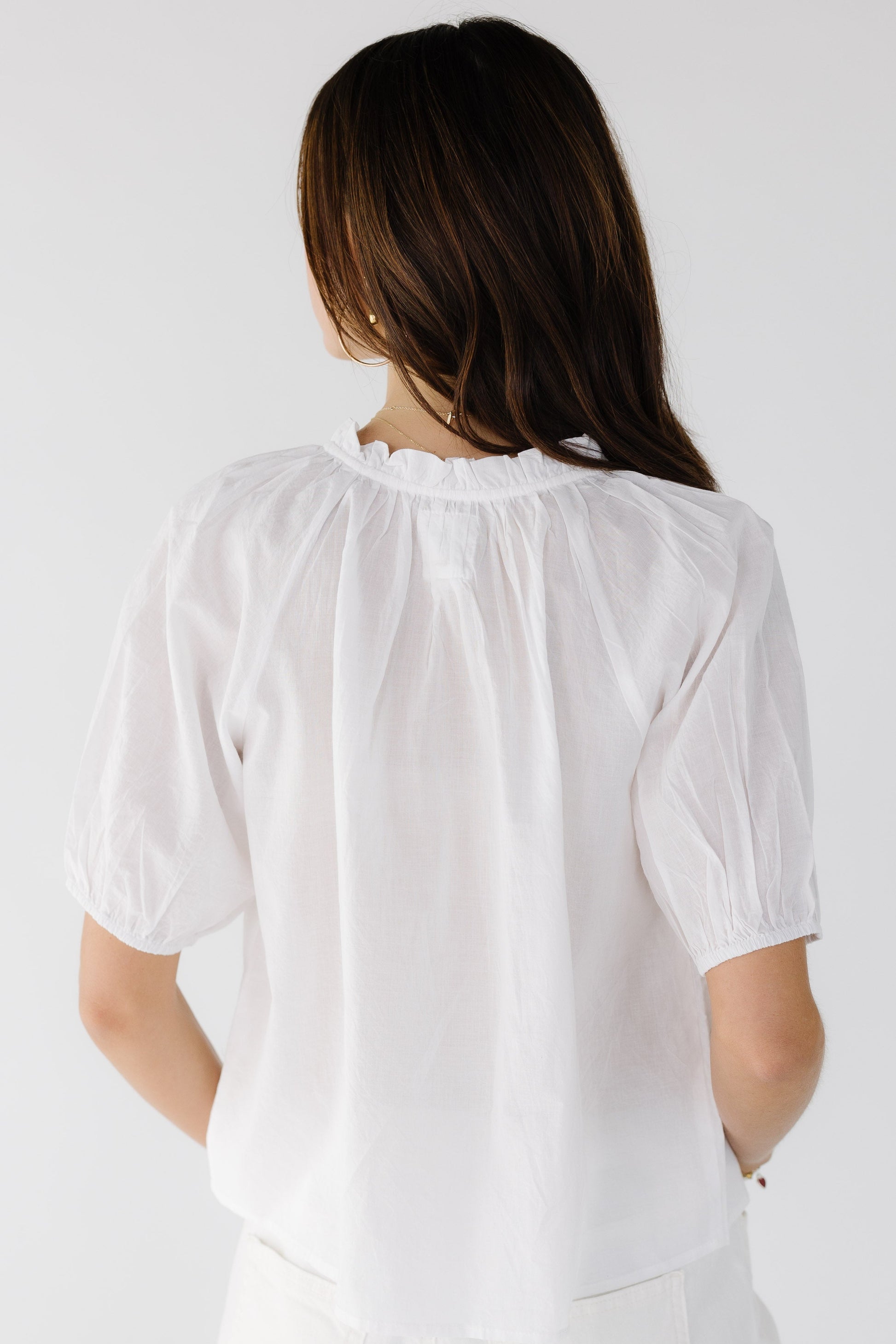 Back view of a young woman wearing a white loose fitting blouse with short gathered sleeves and a ruffled collar.