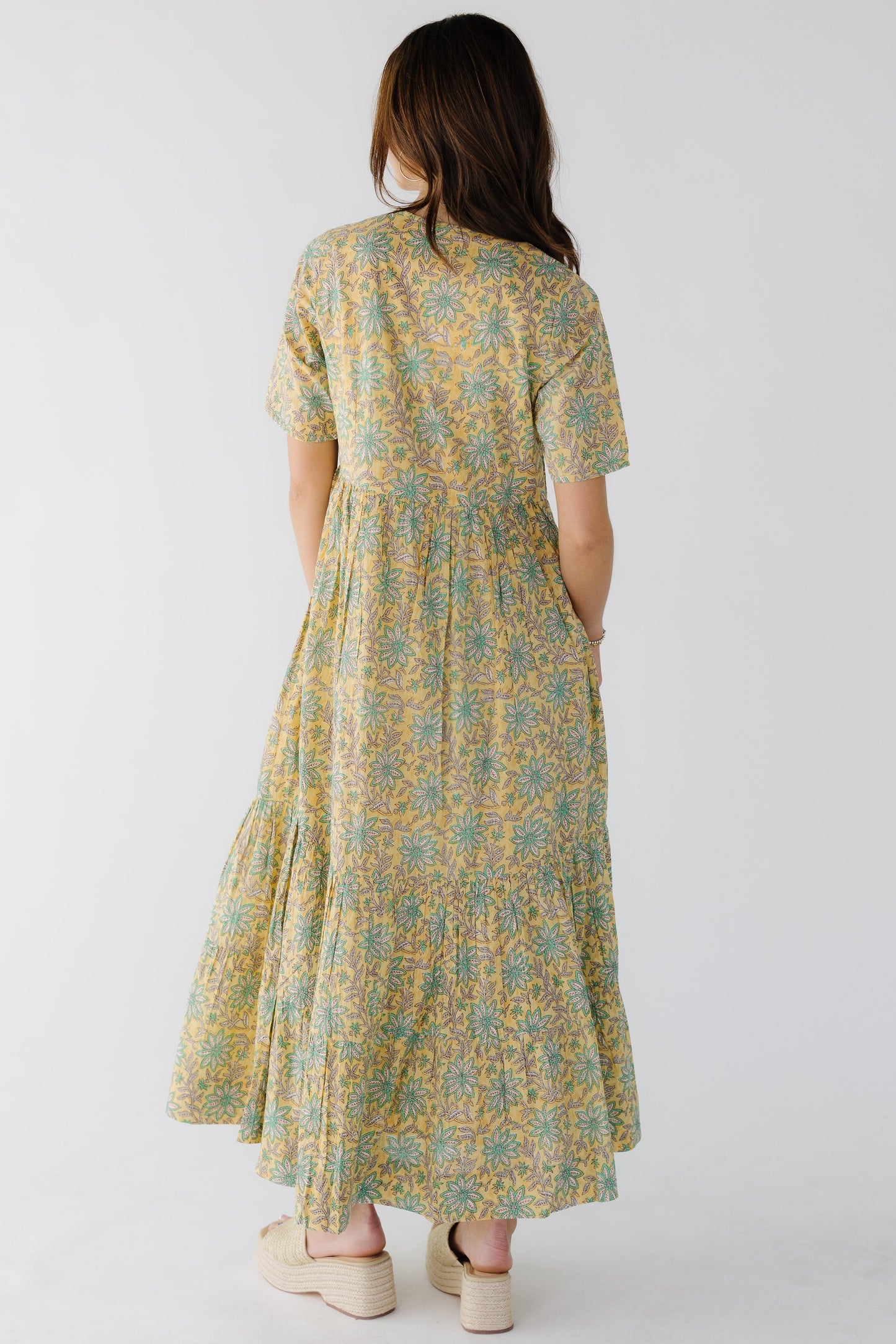Back view of a woman wearing a yellow floral midi dress with short sleeves  full midi skirt with a wide bottom ruffle.