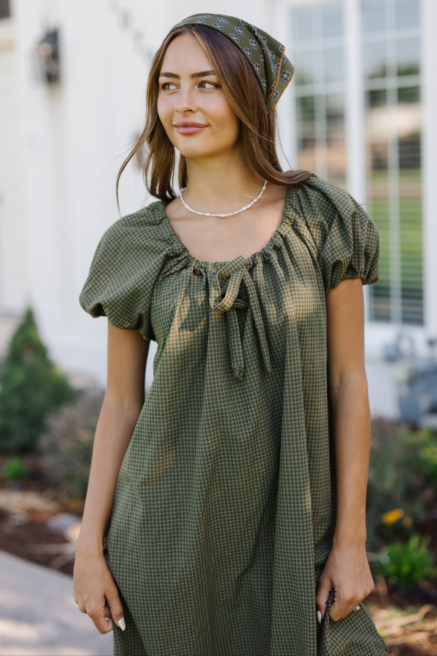 Closer view of a young woman wearing a green gingham, loose fitting dress with puff sleeves and a drawstring-scoop neckline.