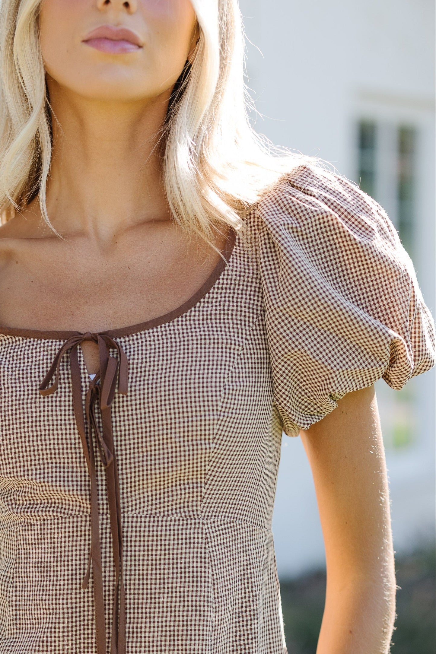 Close view of a woman wearing a brown & cream gingham dress with a wide scoop neckline with 2 front ties, and balloon sleeves.