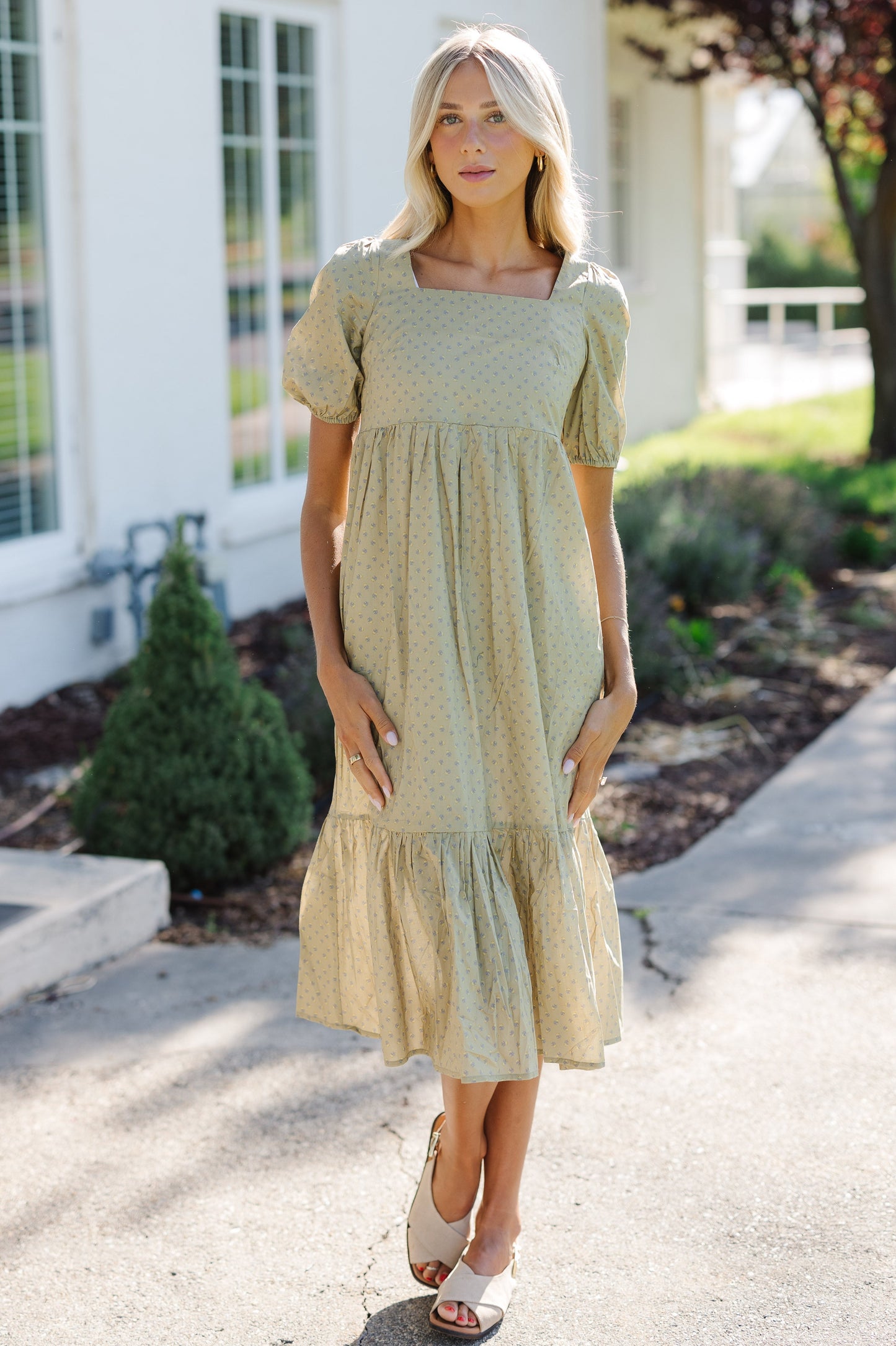 Woman wearing a modest olive print midi dress with a square neckline, large puff sleeves and a wide bottom ruffle.