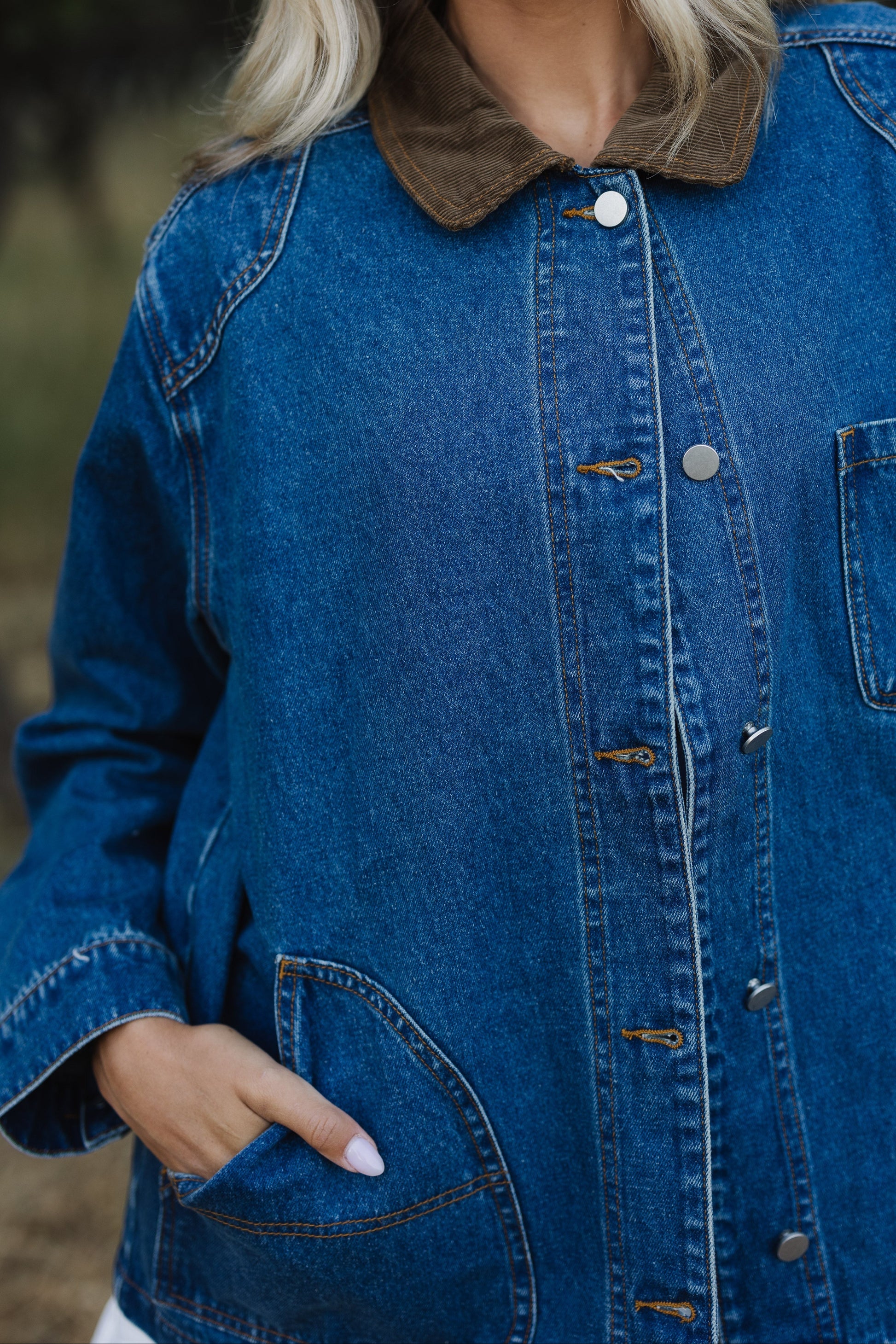 Close view of a longer length denim jacket with  curved front pockets metal buttons and a corduroy pointed collar.