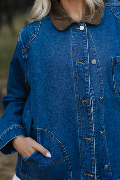 Close view of a longer length denim jacket with  curved front pockets metal buttons and a corduroy pointed collar.