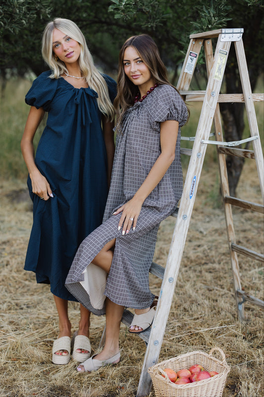 Two young women wearing matching, modest loose fitting midi dress in navy and black plaid with drawstring-scoop necklines and large puff sleeves.