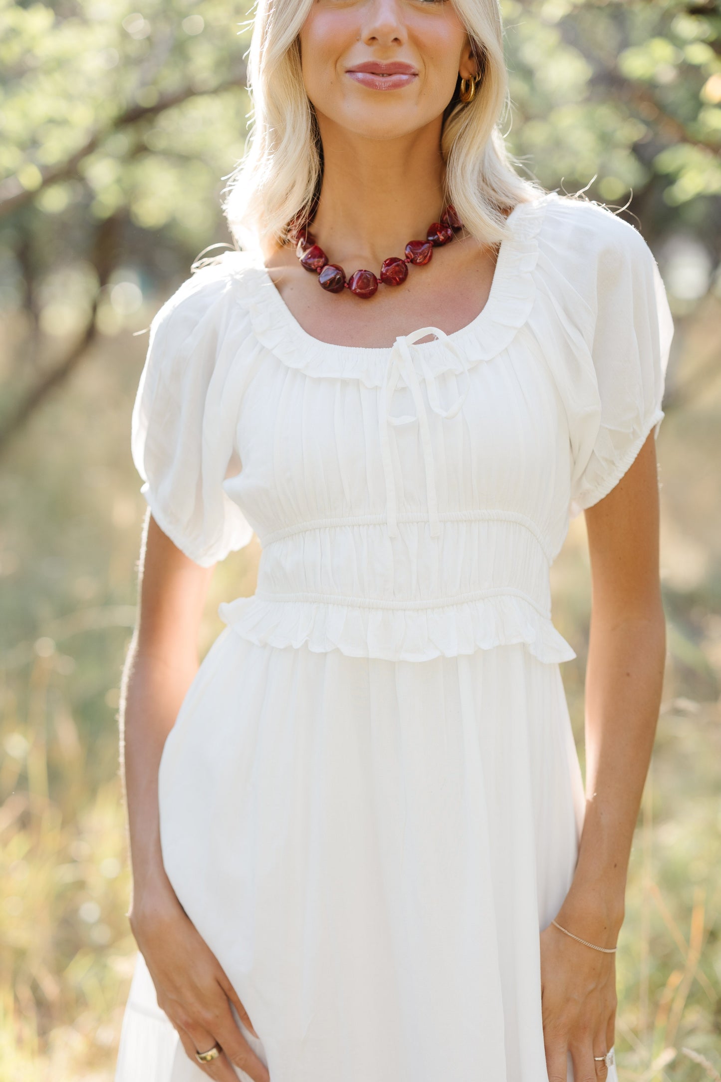 Close up view of a woman wearing a white  dress with a ruffled scoop neckline, short puff sleeves, wide waist with a ruffle and full skirt.